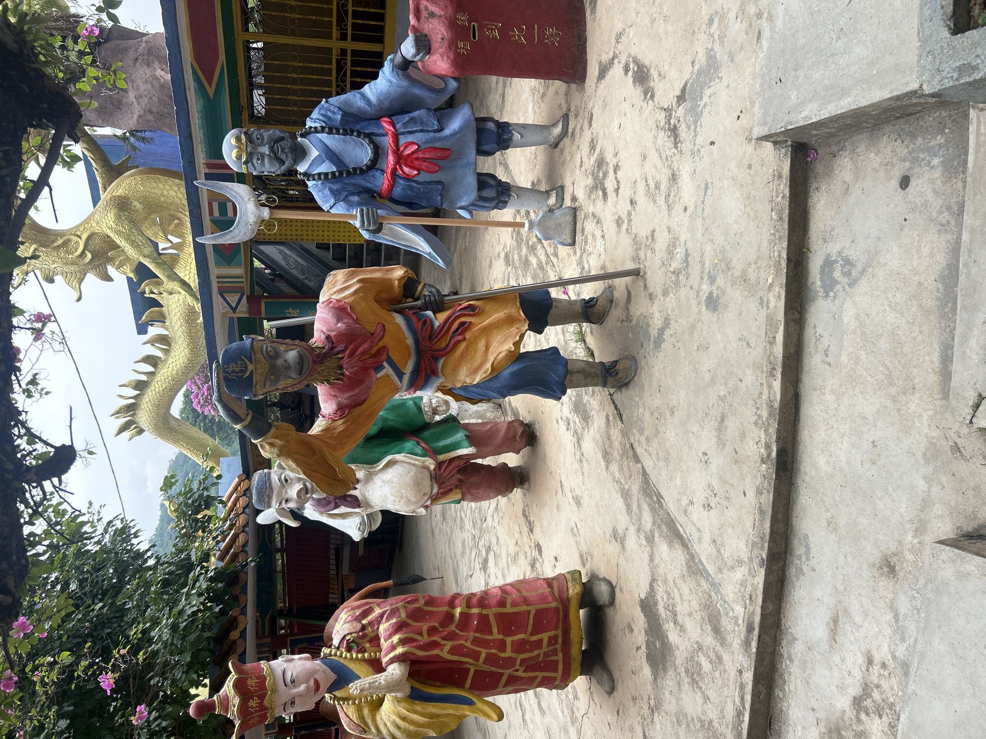 The image depicts a collection of statues in a courtyard, possibly part of a temple or religious site in Ipoh, Malaysia. In the foreground, from left to right, are four statues. The leftmost statue is a figure in a red and gold robe with a tall, ornate headdress, hands clasped in a prayer-like gesture. To its right stands a statue with a pig-like head, wearing a green robe and a white cloth draped over its belly, holding a sack. Next is a statue of a monkey-like character in a bright orange and blue outfit, wearing a blue cap, holding a staff. The rightmost statue is a figure in a blue robe with a red sash and a black beaded necklace, holding a pole with a crescent-shaped tool at the top. Behind these figures, a large, golden dragon sculpture coils above a colorful building. A red monument with Chinese inscriptions stands next to the blue-robed statue. The ground is paved with concrete, showing some wear and discoloration. Trees with pink flowers are visible in the upper left, and a hilly, vegetated landscape is in the background under an overcast sky.