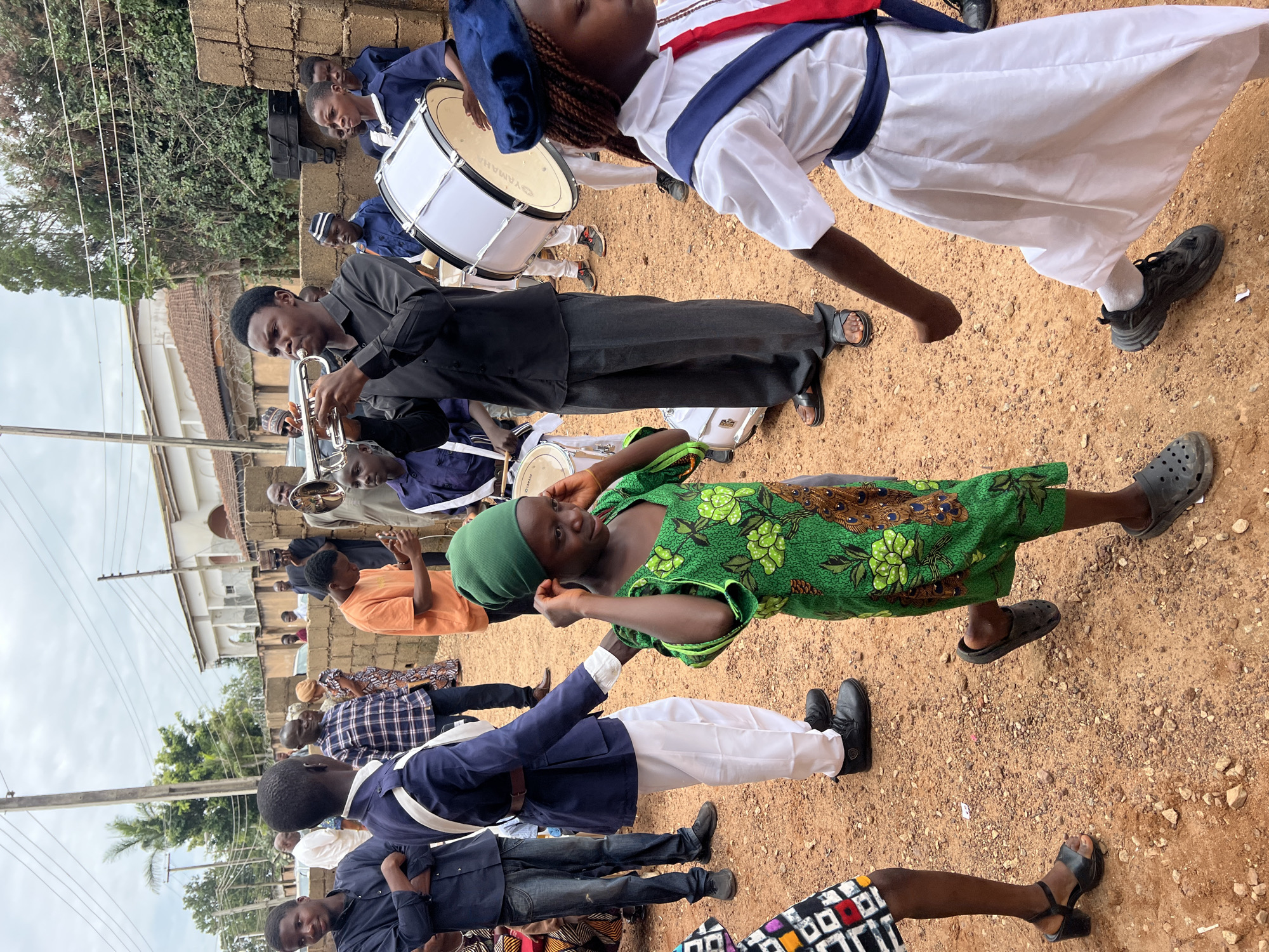 This image depicts a procession or gathering outdoors in a dusty, reddish-brown environment, likely in the city of Jos, Nigeria. The scene is filled with people, many of whom are children and young adults, dressed in a mix of casual and formal attire, with some in what appear to be uniform-like outfits.
In the foreground, a young girl wearing a vibrant green dress with a peacock-feather-like pattern is turning towards the viewer, her hands near her ears, suggesting she might be reacting to music or noise. To her right, another young person, possibly a girl, is wearing a white uniform with a blue sash and a red tie, and a blue beret. Her arm is extended as if to play a musical instrument or steady herself.
Further back, a young man in a black shirt and gray trousers is playing a trumpet. Next to him, a large white bass drum is visible, and to the right, a group of boys in blue uniforms are gathered. One boy is holding a drumstick. In the background, adults are observing the scene, some dressed in traditional Nigerian attire and others in casual wear. Behind the gathering, there is a brick wall and vegetation, with buildings visible in the distance. The sky is overcast. The ground is covered in loose dirt and small stones. The overall atmosphere appears to be celebratory or ceremonial.