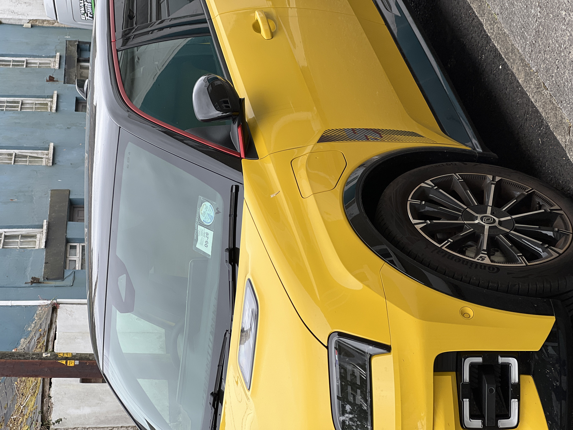 A bright yellow car with a black roof and red trim is parked on a street. The car has distinctive headlights and a textured panel on the side. A dark-colored side mirror is visible. A sticker with "09-26" and "HIGHLAND MOTORS" is on the windshield. In the background, there are buildings with blue and grey facades, some with white window frames. A wooden pole is also present. The street surface is asphalt.