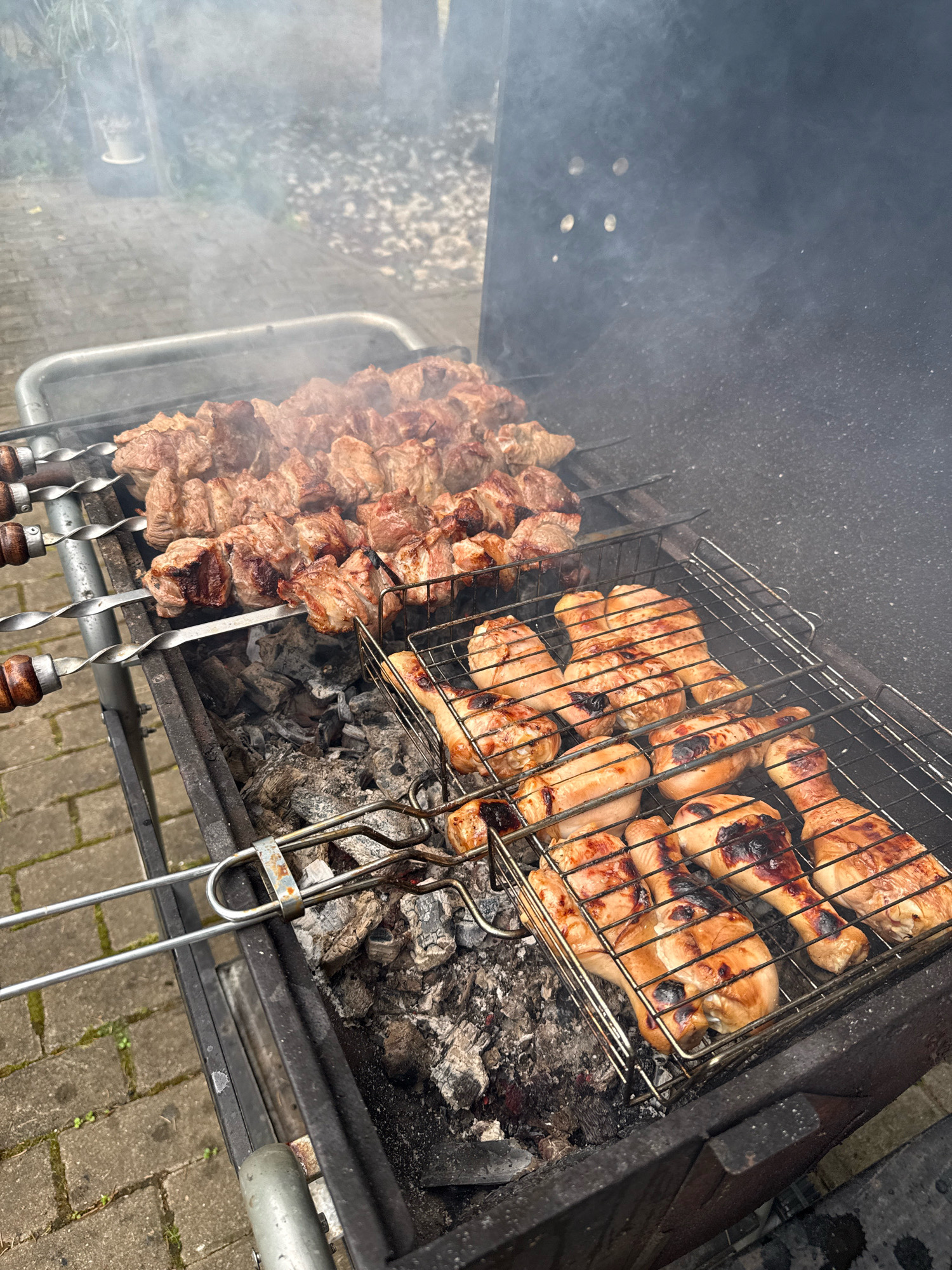 The image shows a close-up view of a barbecue grill with various meats cooking. On skewers, chunks of meat, likely pork, are being grilled. In a wire basket on a lower rack, chicken drumsticks and thighs are cooking. The meats are browned and appear to be charred in places, indicating they are well underway. Smoke rises from the grill, obscuring some of the background. The grill itself is situated outdoors, with a paved surface visible in the foreground, suggesting a patio or outdoor cooking area. The background is blurred but hints at foliage and a building. The scene is consistent with a casual outdoor meal being prepared.