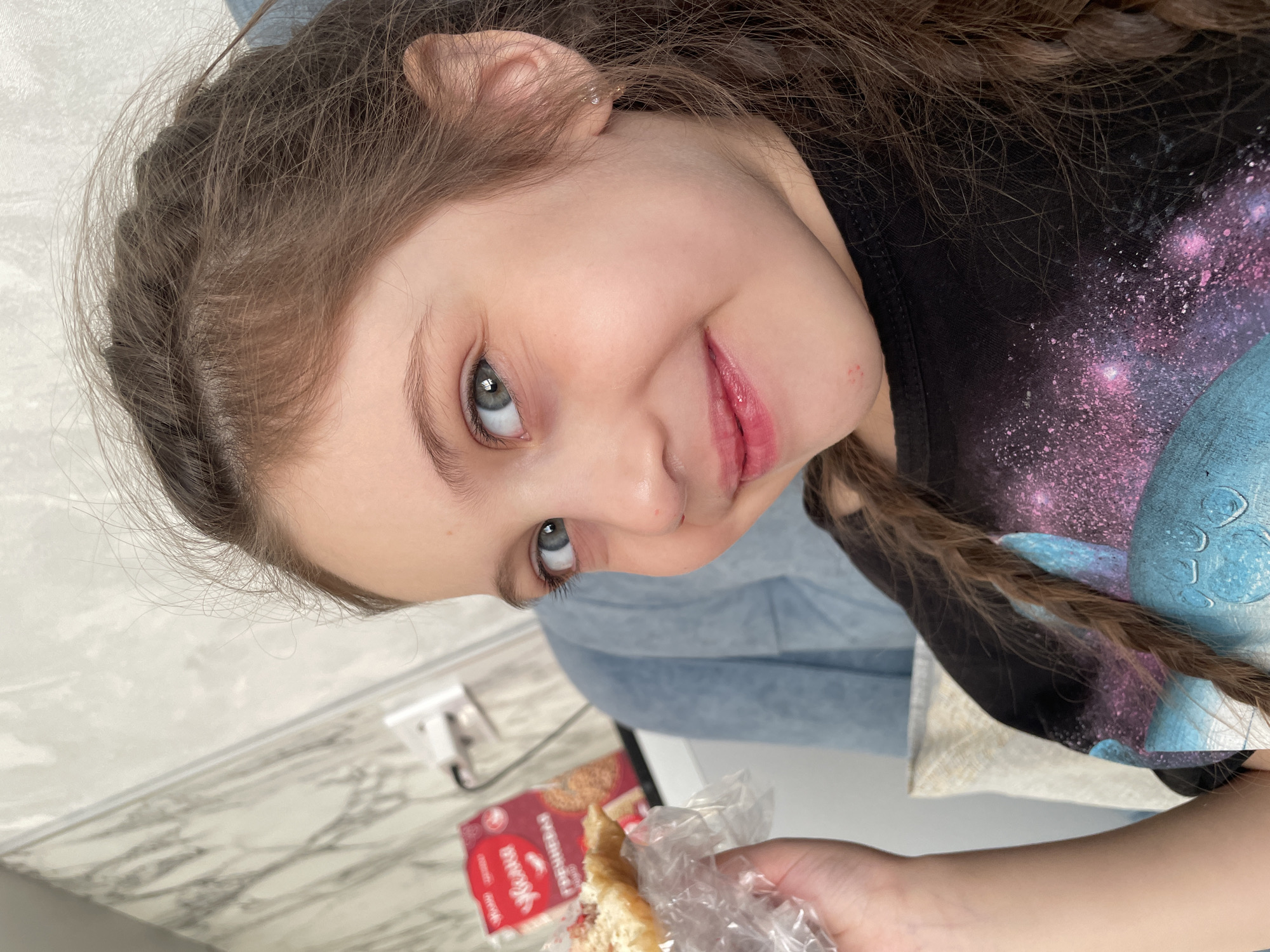 A young girl with light skin and blue eyes is gazing up and smiling while holding a half-eaten sandwich in a plastic wrapper. Her long, brown hair is styled with a braid visible on the right side. She is wearing a black t-shirt with a print of stars and a blue design. In the background, there is a section of what appears to be a light-blue cushioned chair and a white wall or countertop surface with a faux marble pattern. On the counter next to the girl's hand is a red package. Based on available information, it is not possible to confirm the exact location in the city of Izhevsk, Russia.