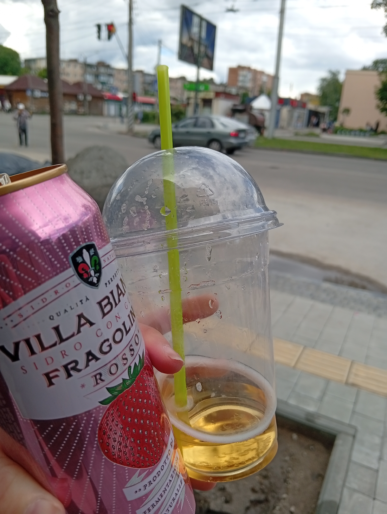 A can of Villa Biance Sidro Fragolino Rosso sits next to a plastic cup with a straw, positioned on a sidewalk in the city of Poltava, Ukraine. The sidewalk is made of square gray tiles and runs along a street with a yellow curb. A street with traffic, including a gray car, is visible in the background. Buildings and a traffic light can be seen across the street. A blurry figure of a person can be seen further in the background. The sky appears overcast.
