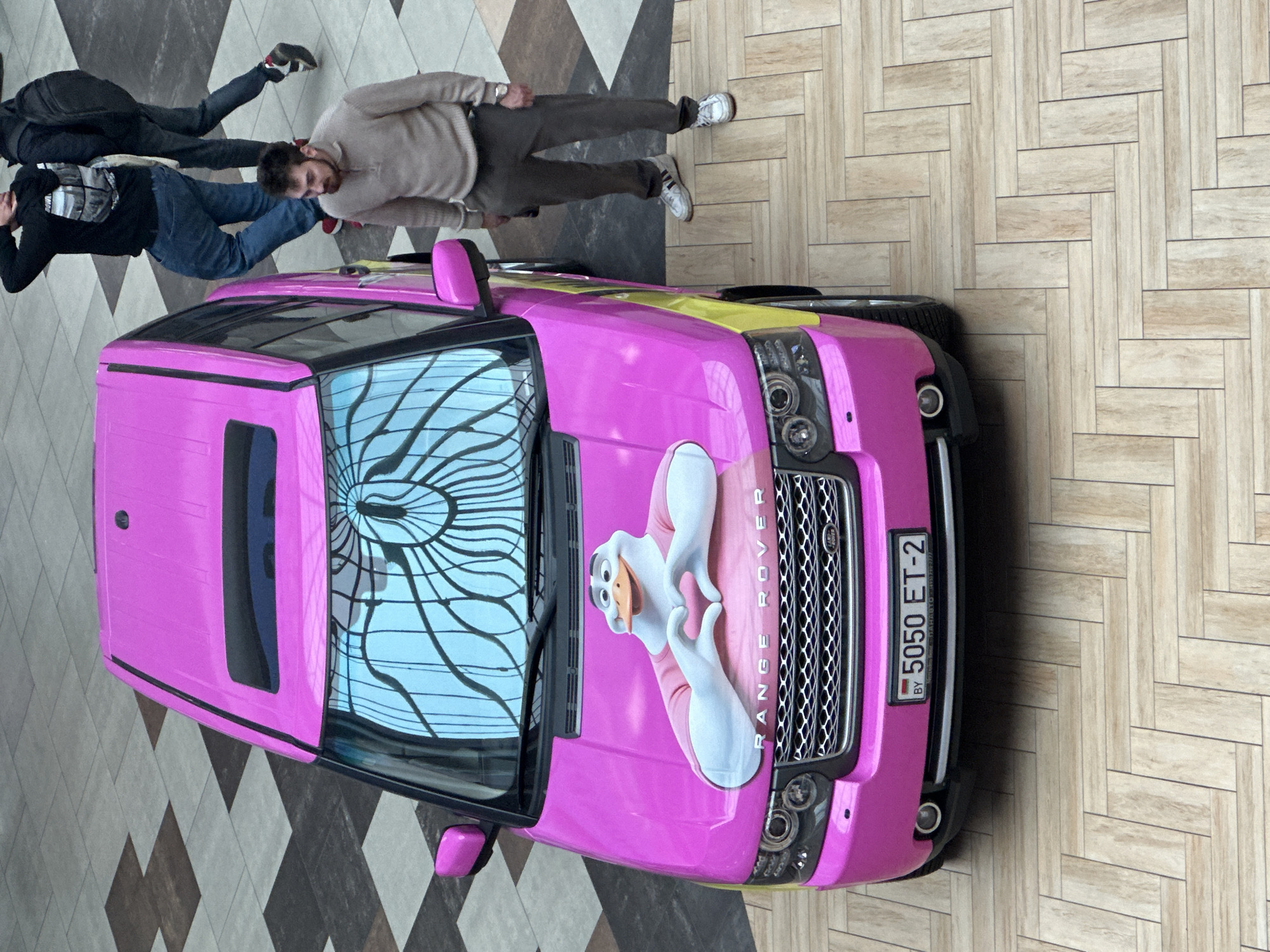 The image depicts a top-down view of a pink Range Rover SUV parked on a herringbone patterned wooden floor, likely inside a shopping mall or similar indoor public space in Minsk, Belarus. The car is adorned with a large decal of a cartoon duck character on its hood, making a heart shape with its hands. The license plate of the car reads "5050 ET-2 BY".
Two men are visible in the background, walking away from the car. One man, wearing a black hooded sweatshirt and blue jeans, has his hand raised to the back of his head. The other man, dressed in dark trousers and a light-colored sweater, is looking down towards his feet. Both men are walking on a floor covered in a checkered pattern of grey and brown tiles. The overall lighting suggests an indoor environment.