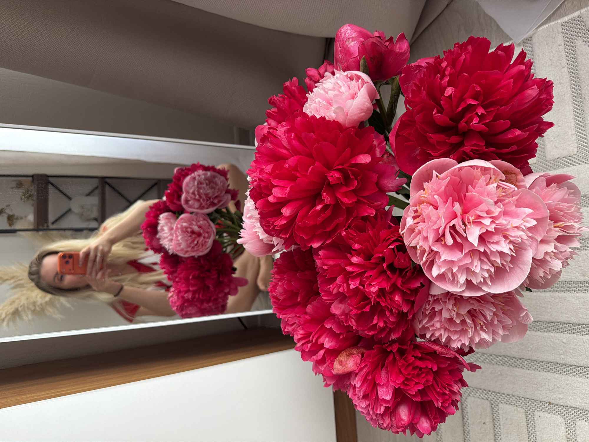 A woman is reflected in a mirror, holding a phone up to take a selfie. She is wearing a red and white striped shirt. In the foreground, a bouquet of bright pink and fuchsia peonies sits in a vase. The background of the mirror reflection shows a shelf unit with decorative items and some dried pampas grass. The setting appears to be an indoor residential space. There are no specific indicators in the image to suggest a location in Moscow, Russia.