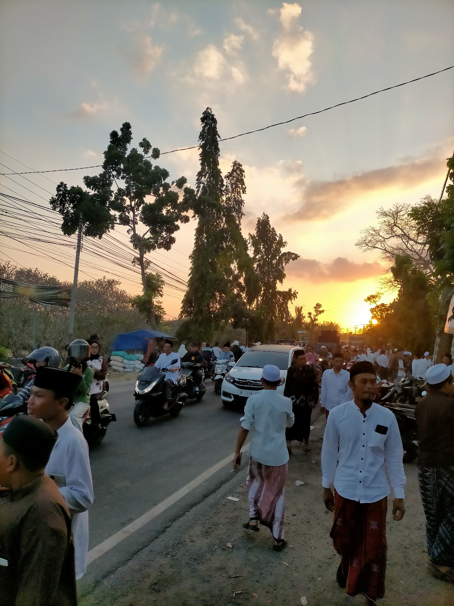 An outdoor, eye-level, medium shot captures a bustling street scene in Kranji, Indonesia, during sunset. The sky is ablaze with warm, orange and yellow hues, transitioning to pale blue as it moves upwards, with scattered white clouds. Silhouetted against this vibrant sky are tall, slender trees on the left and a more spread-out tree canopy on the right, partially obscuring the horizon. Electrical wires crisscross the sky.
The foreground is dominated by a paved road with a white line marking its edge. Several people, mostly men dressed in white shirts and sarongs, and a few women in headscarves, are present. Some are walking on the side of the road, while others are on or around motorcycles.
On the road, several motorcycles are visible, some with riders. A white car is positioned in the middle distance, also on the road. People are walking in both directions on the road and along its shoulder. In the background, more people, trees, and a blue tarp covering what appears to be a pile of sandbags are discernible. The overall impression is one of a lively gathering or procession at dusk.
