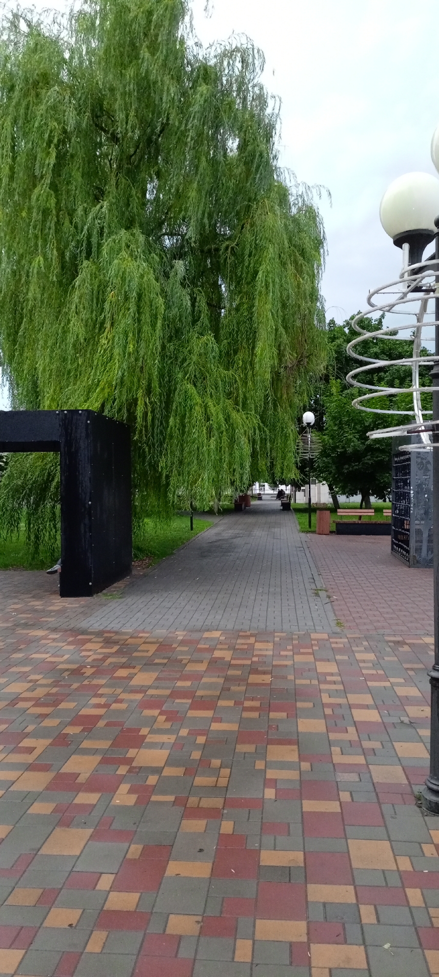 The image depicts a public walkway in the city of Kirsanov, Russia, on an overcast day. The foreground features a paved path with a mosaic of red, tan, and gray rectangular pavers. To the left, a large weeping willow tree with dense green foliage extends over the walkway, partially obscuring a tall, black, rectangular structure. Beyond this, the walkway continues, lined with grass and trees. In the middle ground, a few benches and trash receptacles are visible along the path. Further in the distance, a few individuals are seated on benches, and a lamppost with a spherical light fixture stands to the right. The overall scene suggests a park or a landscaped public area.