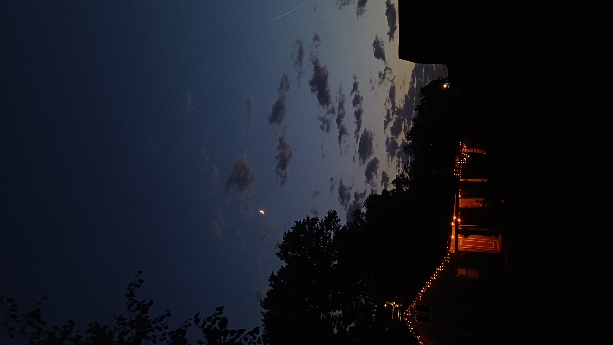 The image shows a nighttime scene with a crescent moon and a planet (likely Venus) visible in the dark blue sky. Several dark clouds are scattered across the sky. Silhouetted trees are visible in the foreground. A building with orange lights is present on the right side of the image. String lights are hung on the building. The image appears to be taken outdoors in Timokhovo, Russia.