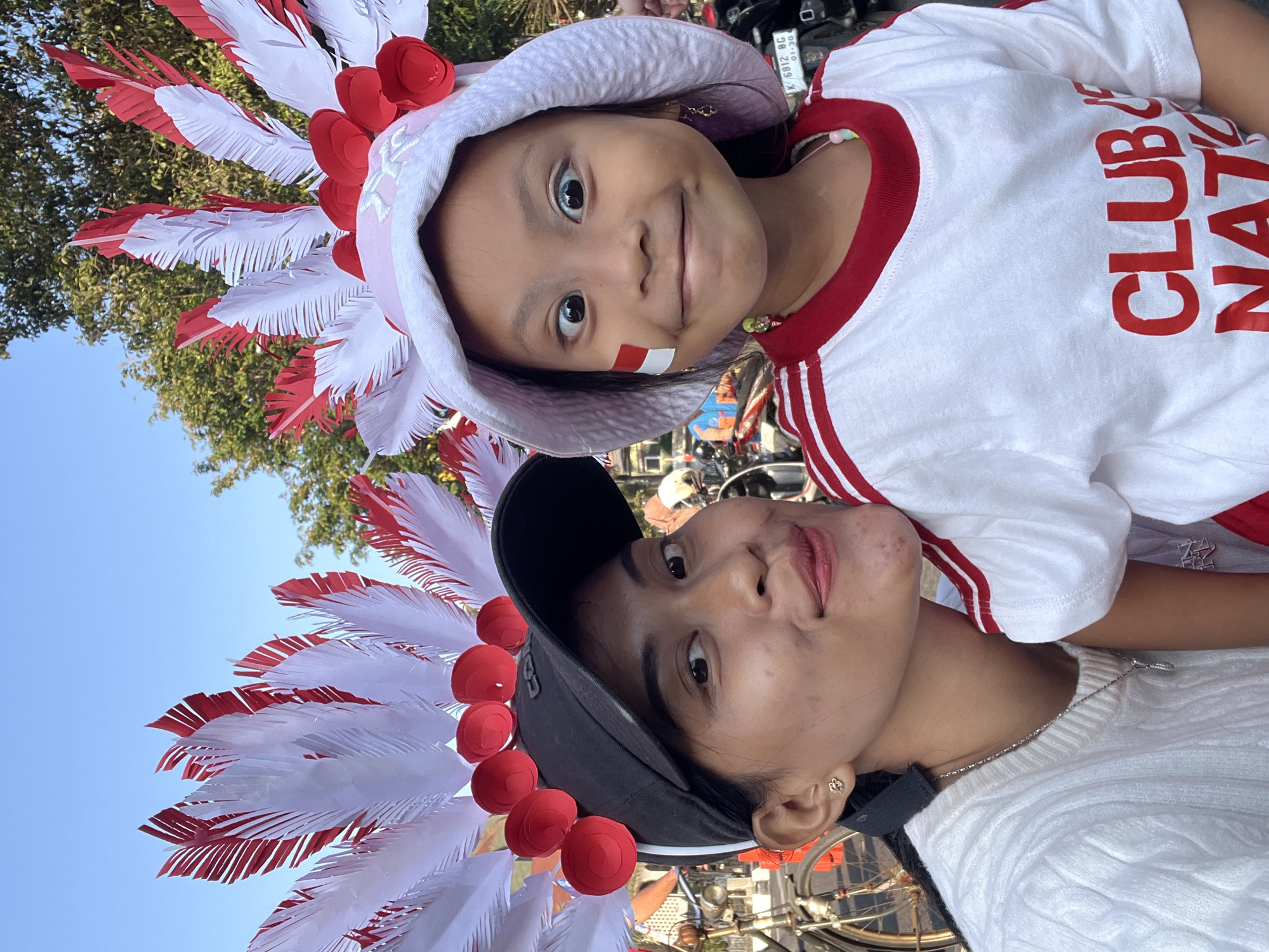 Two individuals, an adult female and a young girl, are captured in a close-up outdoor photograph, likely in Kecamatan Pati, Indonesia. The adult, wearing a white cable-knit sweater and a black baseball cap adorned with red paper rosettes and white and red feathers, faces the camera with a slight smile. The young girl, positioned slightly to the right and in front of the adult, wears a white t-shirt with red stripes on the collar and sleeves, a light pink bucket hat decorated with similar red rosettes and feathered headwear, and has a small Indonesian flag sticker on her cheek. Her eyes are wide and looking in different directions, creating a playful expression. The background suggests an urban setting, with trees and what appears to be a motorcycle partially visible on the right side of the frame. The lighting indicates it is daytime, possibly late afternoon given the warm tones.