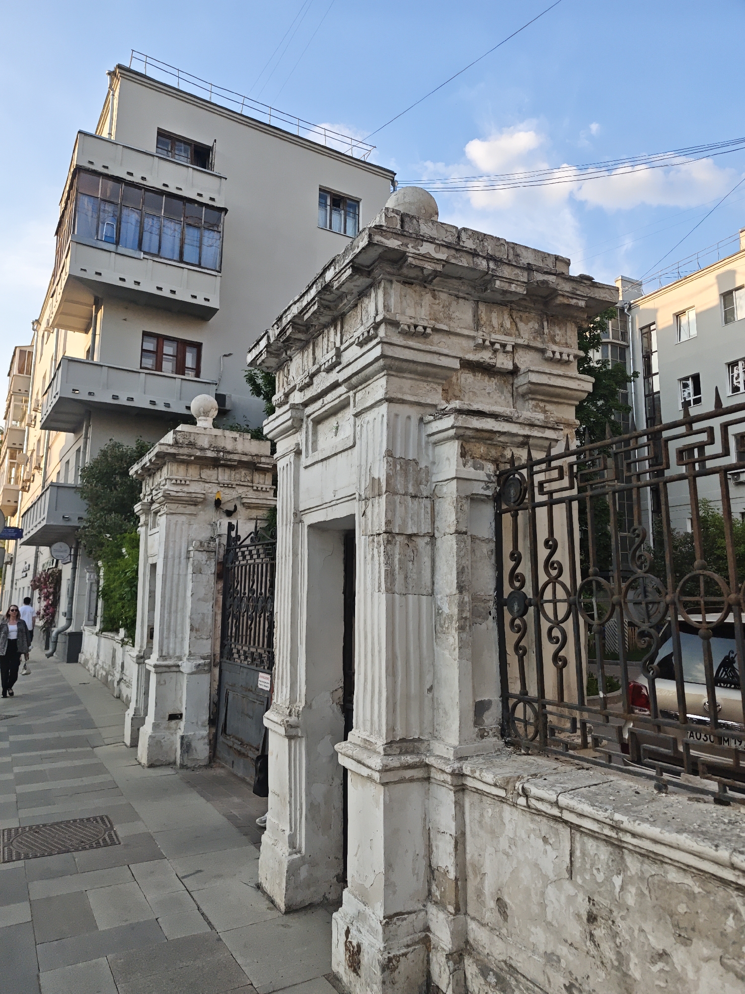 A street view in an unidentified urban location features a sidewalk on the left with a woman in a dark jacket walking away from the camera. The architecture includes a building with a prominent enclosed balcony and a series of columns and ornate metal fencing, which likely encloses a private area. The buildings are light-colored and have a mix of window styles. The sky is blue with scattered clouds. The overall lighting suggests late afternoon or early morning. A car is partially visible through the fence. The infrastructure also includes above-ground cables stretching across the sky. The sidewalk is constructed of rectangular pavers, and there is a drain or utility access cover on the left edge of the frame.