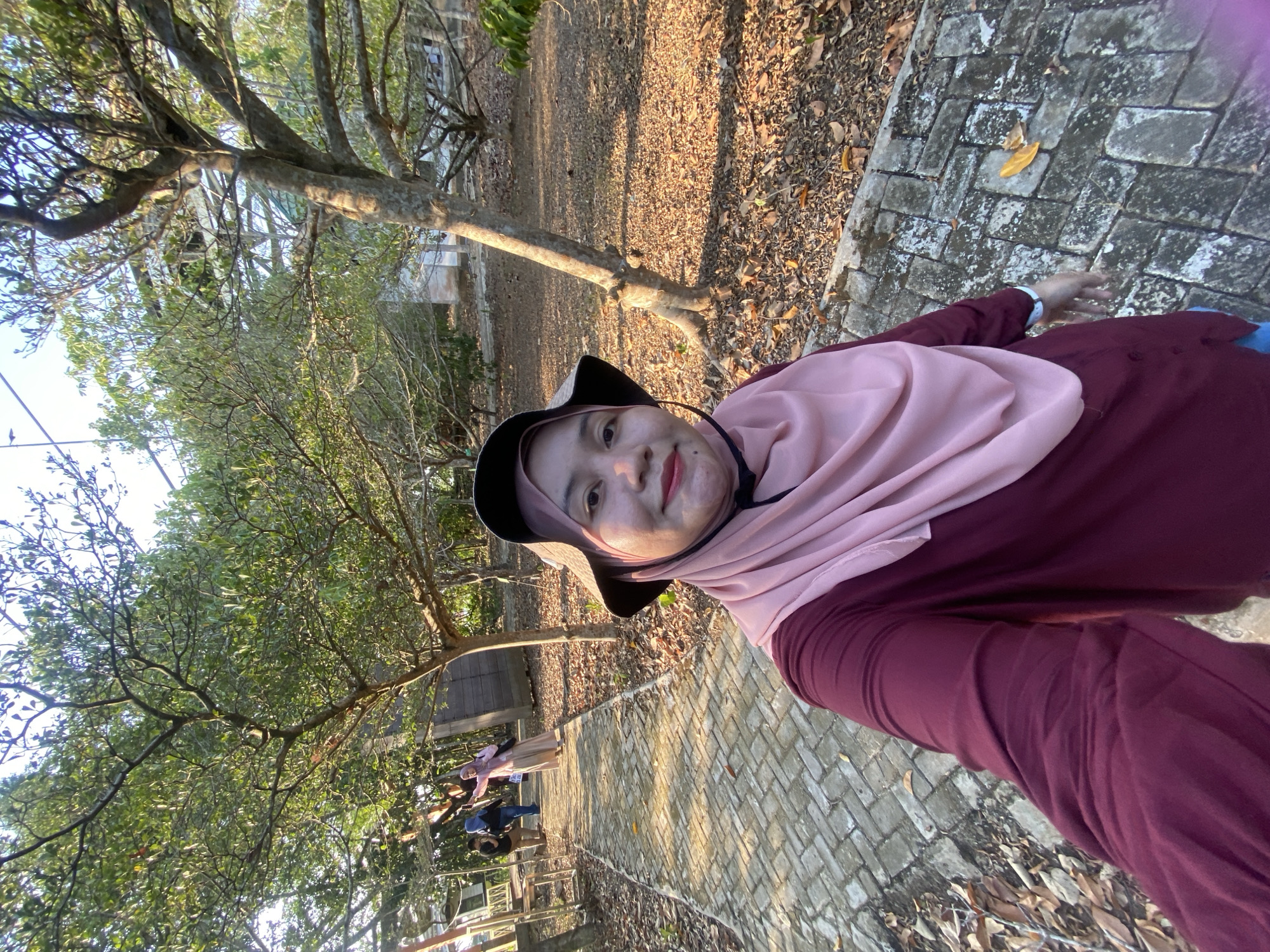 In the city of Sungai Pitung, Indonesia, a woman wearing a maroon shirt, a pink hijab, and a wide-brimmed hat is taking a selfie. She is standing on a brick pathway in an outdoor area with trees and fallen leaves. In the background, several other people, dressed casually, are walking away from the camera down the pathway. The scene appears to be a park or garden, with sunlight filtering through the trees, casting shadows on the ground.