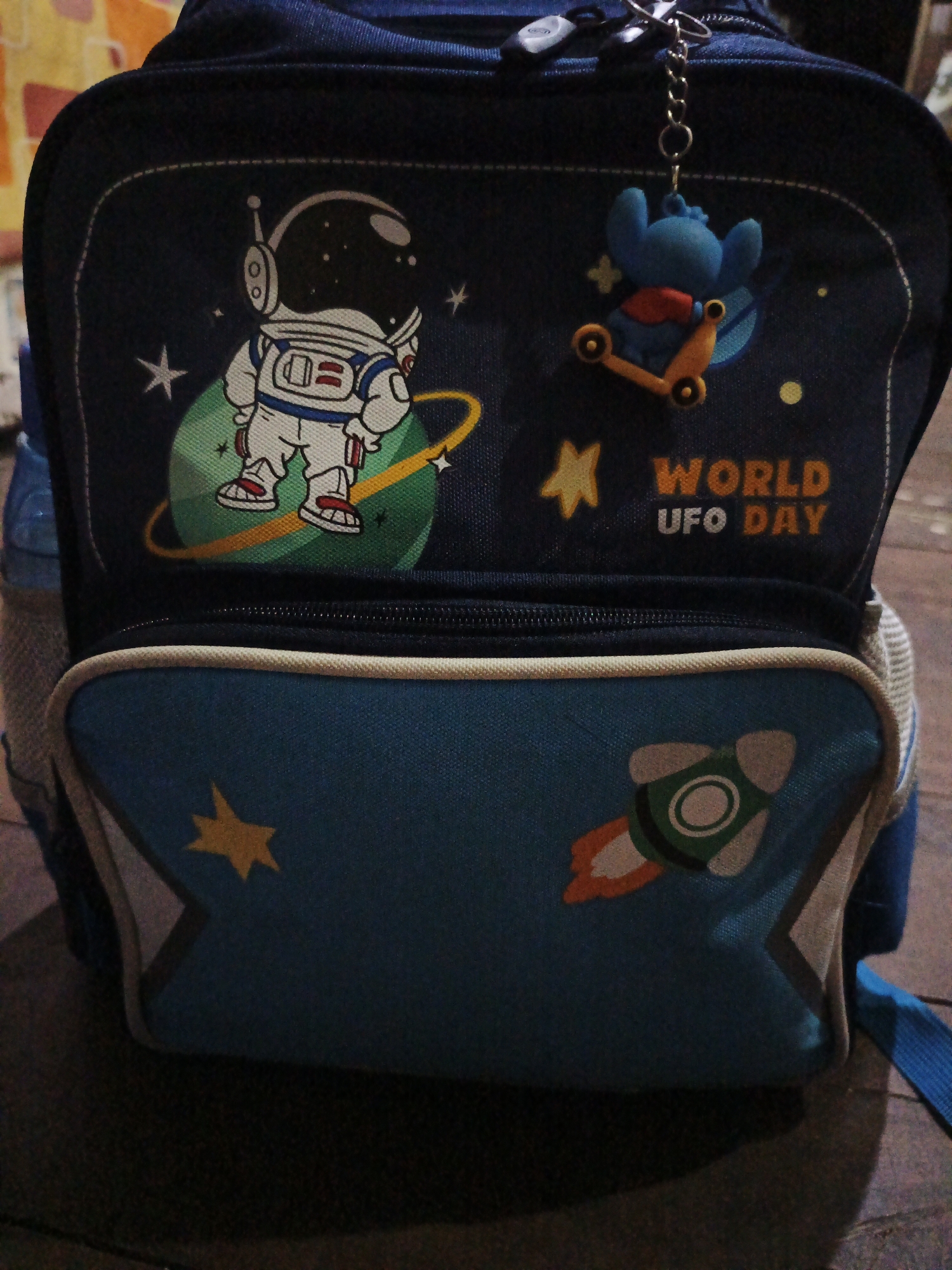 The image shows a close-up, eye-level view of a dark blue children's backpack. The backpack features a colorful graphic design with an astronaut standing on a green planet with a yellow ring. In the background are white stars and yellow dots. A blue cartoon character is attached to the top of the backpack as a keychain, riding a scooter. To the right of the astronaut graphic, the words "WORLD UFO DAY" are printed in orange and white. The lower portion of the backpack has a design of a rocket ship launching with orange flames. The backpack is dark and appears to be indoors with a blurry, textured background that suggests it is against a wall or possibly another bag. The lighting is dim. The image is not specific to Sinar Bulan, Indonesia, or any other particular city.