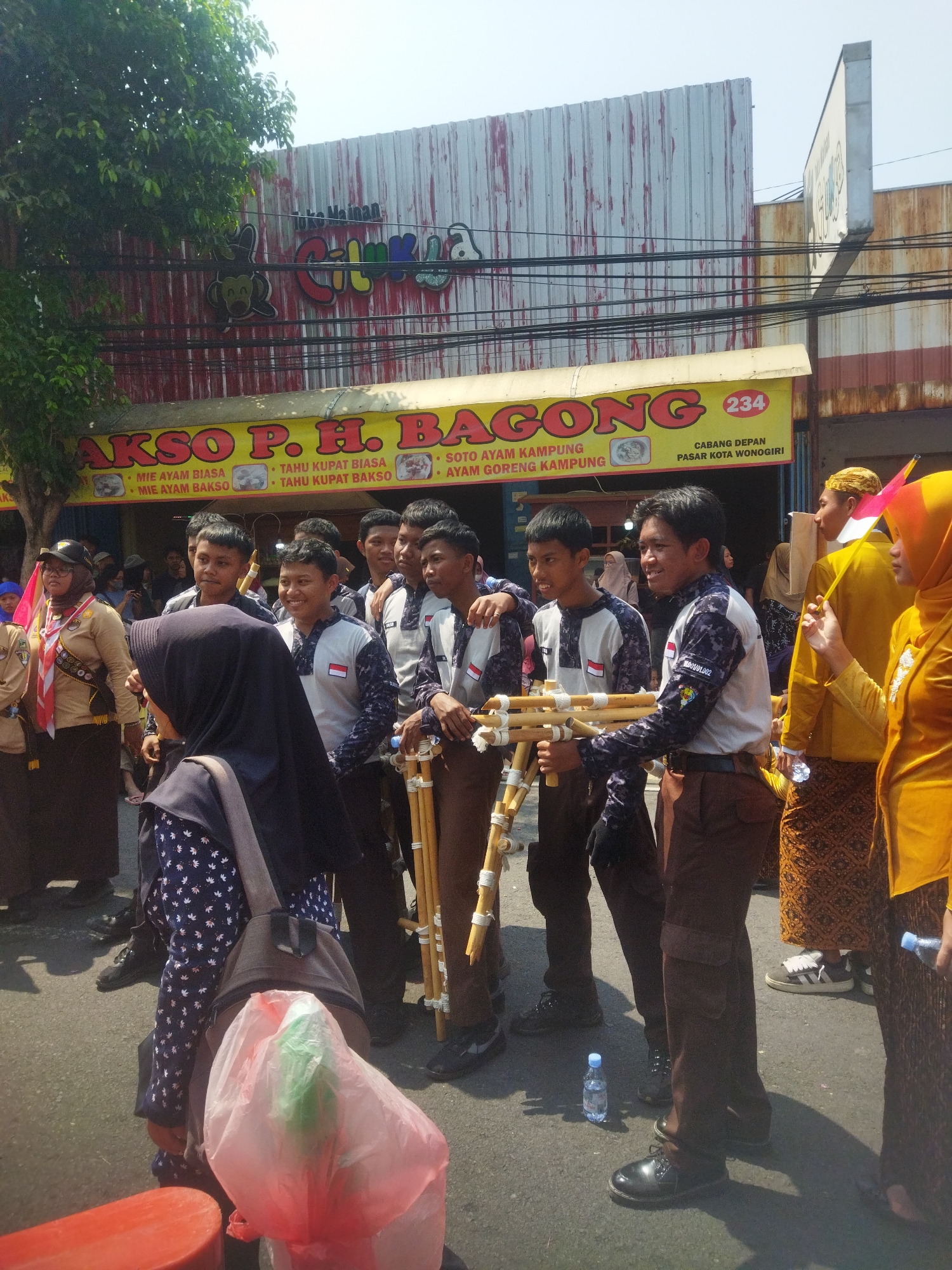 The image depicts a street scene in front of a building in Wonogiri, Indonesia. The building has a corrugated metal facade and a prominent yellow awning with red lettering that reads "AKSO P.H. BAGONG". Below this, the awning lists menu items such as "MIE AYAM BIASA", "TAHU KUPAT BIASA", "SOTO AYAM KAMPUNG", and "MIE AYAM BAKSO". A sign on the awning also indicates "CABANG DEPAN PASAR KOTA WONOGIRI", which translates to "Branch in front of Wonogiri City Market".
In the foreground, a group of young people, likely students, are gathered. Some are wearing scout-like uniforms with khaki shirts and brown pants, while others are in matching gray and white shirts with dark patterned sleeves and brown pants. They appear to be holding bamboo poles bundled together. One young woman in the foreground is wearing a dark hijab and a patterned blue top, carrying a backpack and a pink plastic bag. To the right, two young women are visible, one in a yellow hijab and the other in a yellow top and patterned skirt. The ground is asphalt, and sunlight casts shadows. A small blue water bottle is on the ground near the group. The overall atmosphere suggests a local event or gathering.