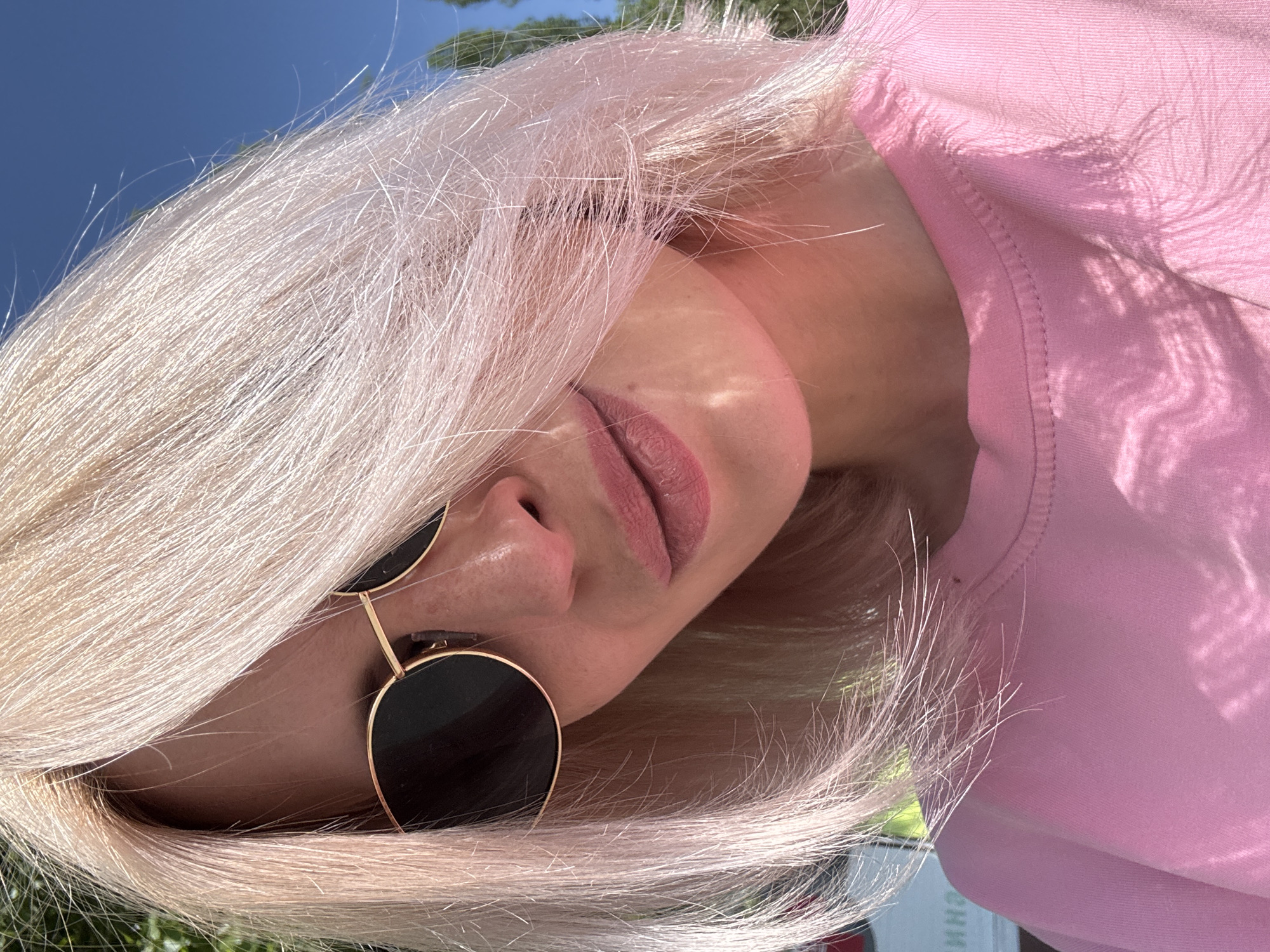 A woman with fair skin and blonde hair partially obscuring her face is wearing round-framed sunglasses and a pink top. Her face is angled towards the camera. The background is a mix of foliage, blue sky and what appears to be a building. The photo appears to be taken outdoors in daylight, based on the sunlight and shadows. The location cannot be accurately determined as Saratov, Russia, since there are no contextual clues from the image to indicate it is in this specific location.