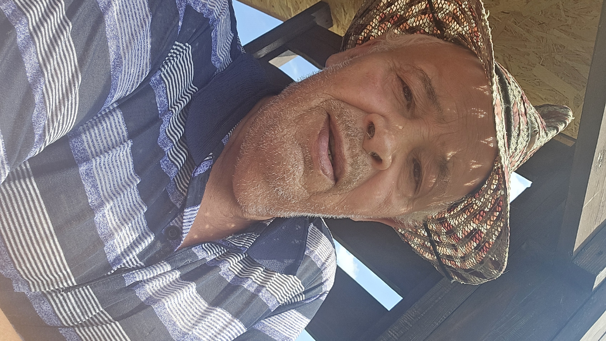 A close-up selfie shows an elderly man wearing a straw hat and a blue and white striped shirt. He is outdoors, with a wooden structure and the sky visible in the background. The man has light skin, gray hair, and a light beard. He is looking directly at the camera with his mouth slightly open. The image appears to have been taken during the day, with sunlight casting shadows on the man's face and hat. The location is not determinable from the image itself.