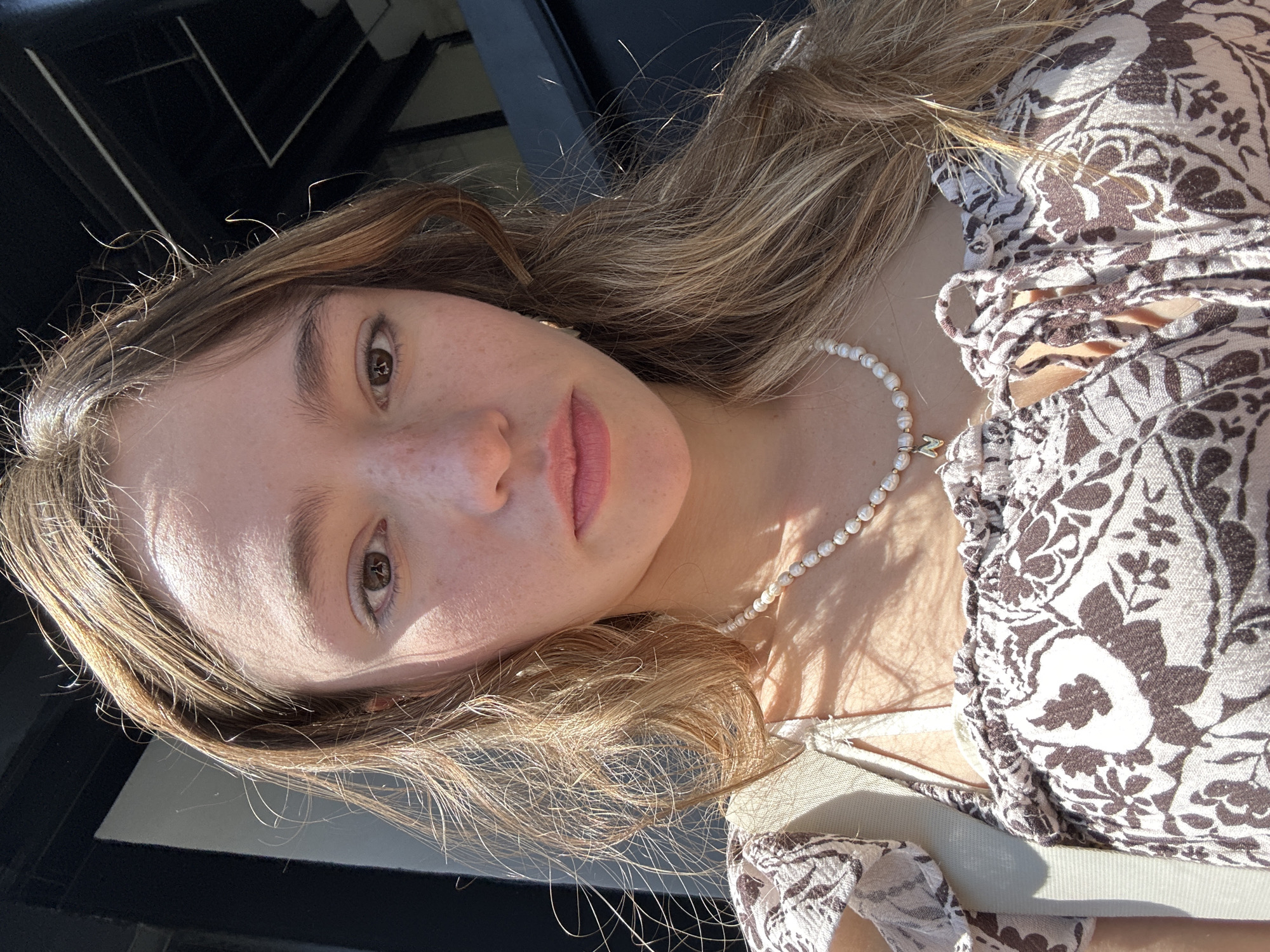 A young woman with blonde hair and freckles is taking a selfie outdoors. She is wearing a brown and white floral print top with a ruffled neckline and a beaded necklace with a gold letter 'N' charm. The background shows a dark doorway or structure with some bright light. She appears to be in the city of San Bernardino Tlaxcalancingo, Mexico.