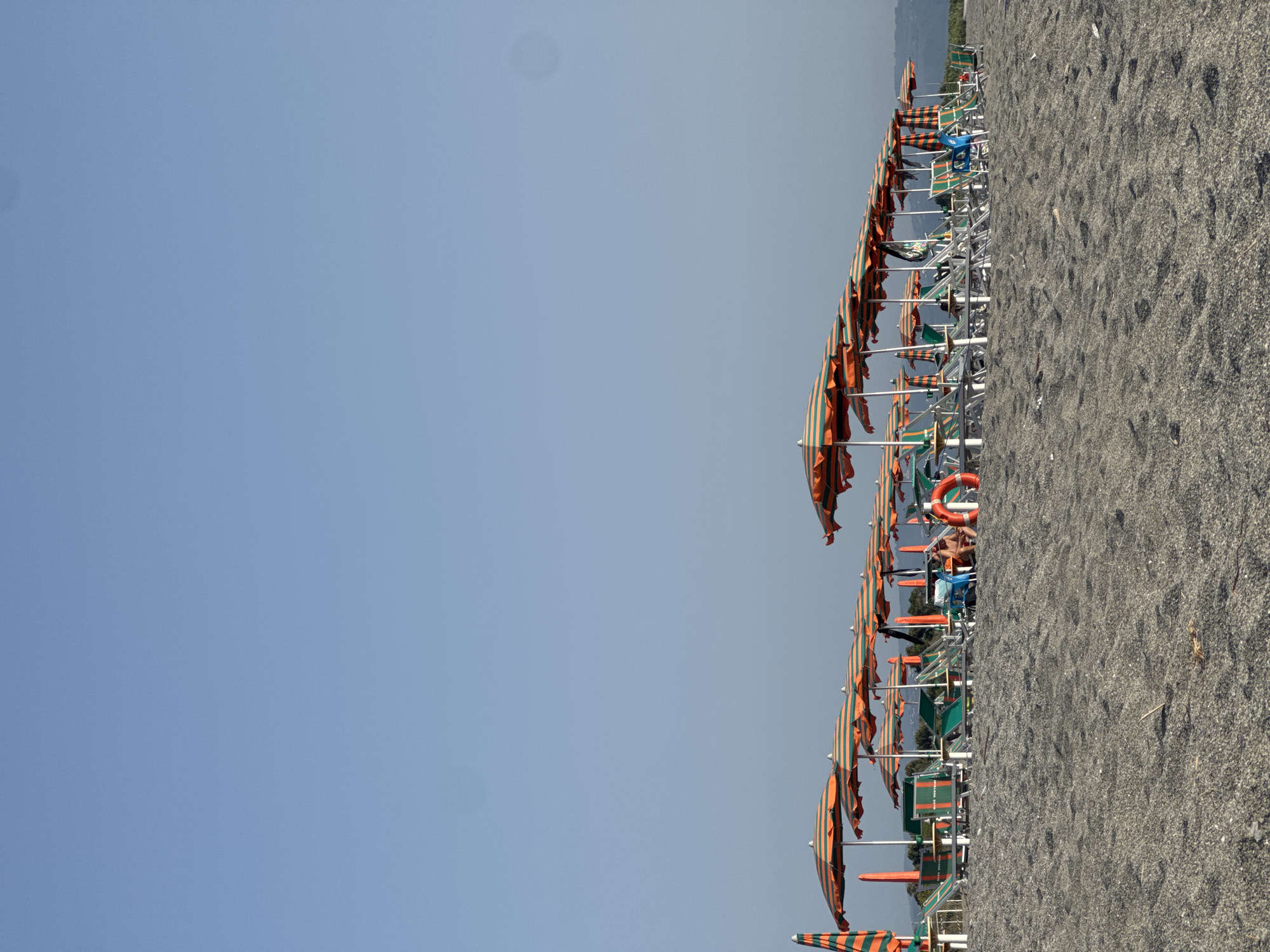 The image depicts a beach lined with numerous orange and green striped umbrellas and lounge chairs. The beach is covered in dark, coarse sand. In the background, there are some trees and hills visible under a clear, pale blue sky. A few people are seated on lounge chairs, and a life preserver is visible. The scene appears to be a resort area in Lamezia Terme, Italy.