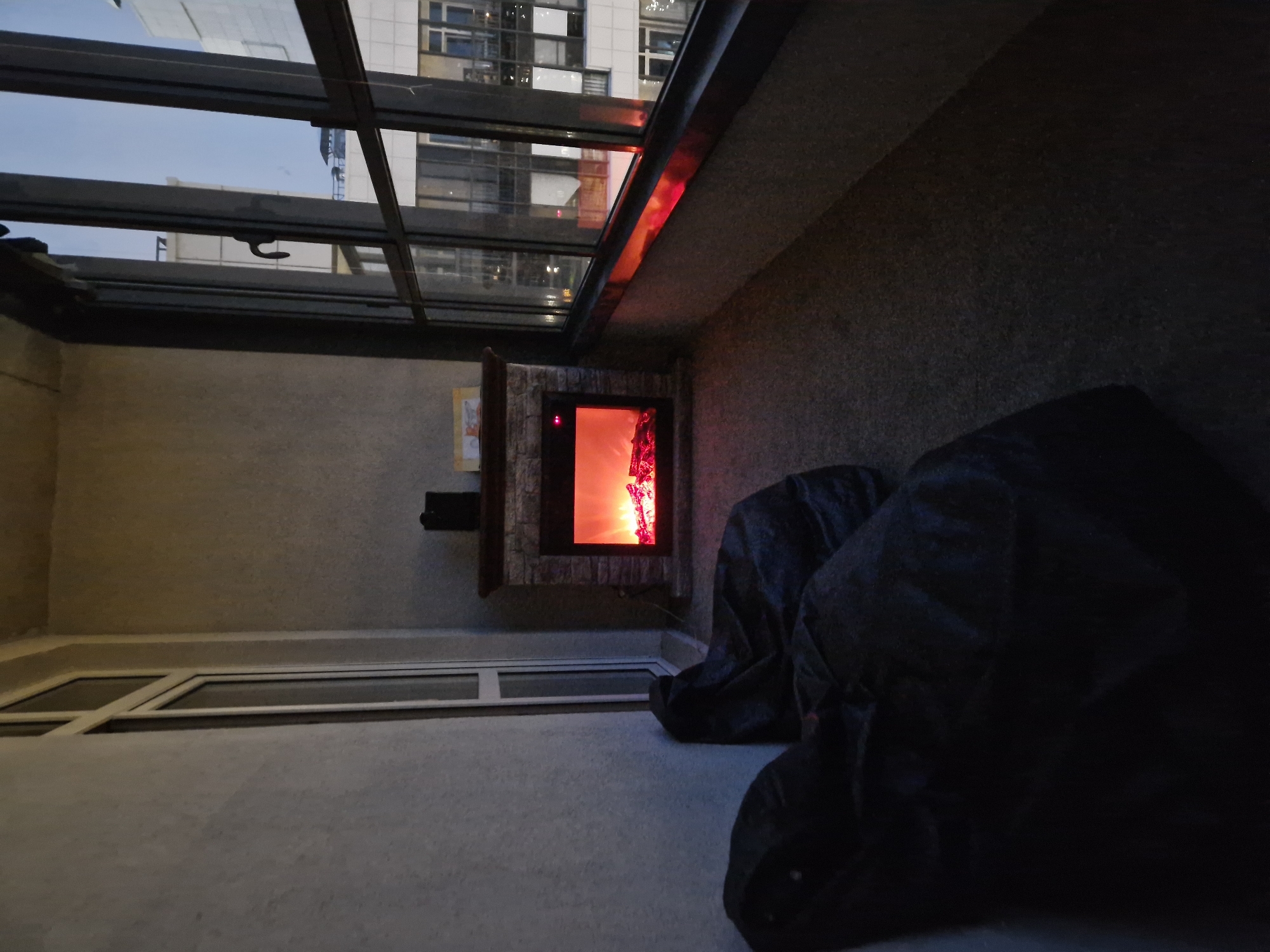 The image depicts a cozy balcony area with an electric fireplace. Two dark-colored beanbag chairs are positioned on the left side of the balcony, facing the fireplace. The fireplace, made of stone and wood, is illuminated with a realistic flame effect, casting a warm glow. Above the fireplace, a small framed picture is visible. The balcony features large windows that offer a view of city buildings. The evening sky is visible through the windows, suggesting it is twilight. The floor of the balcony is covered with a textured carpet. The overall atmosphere is one of comfort and relaxation.