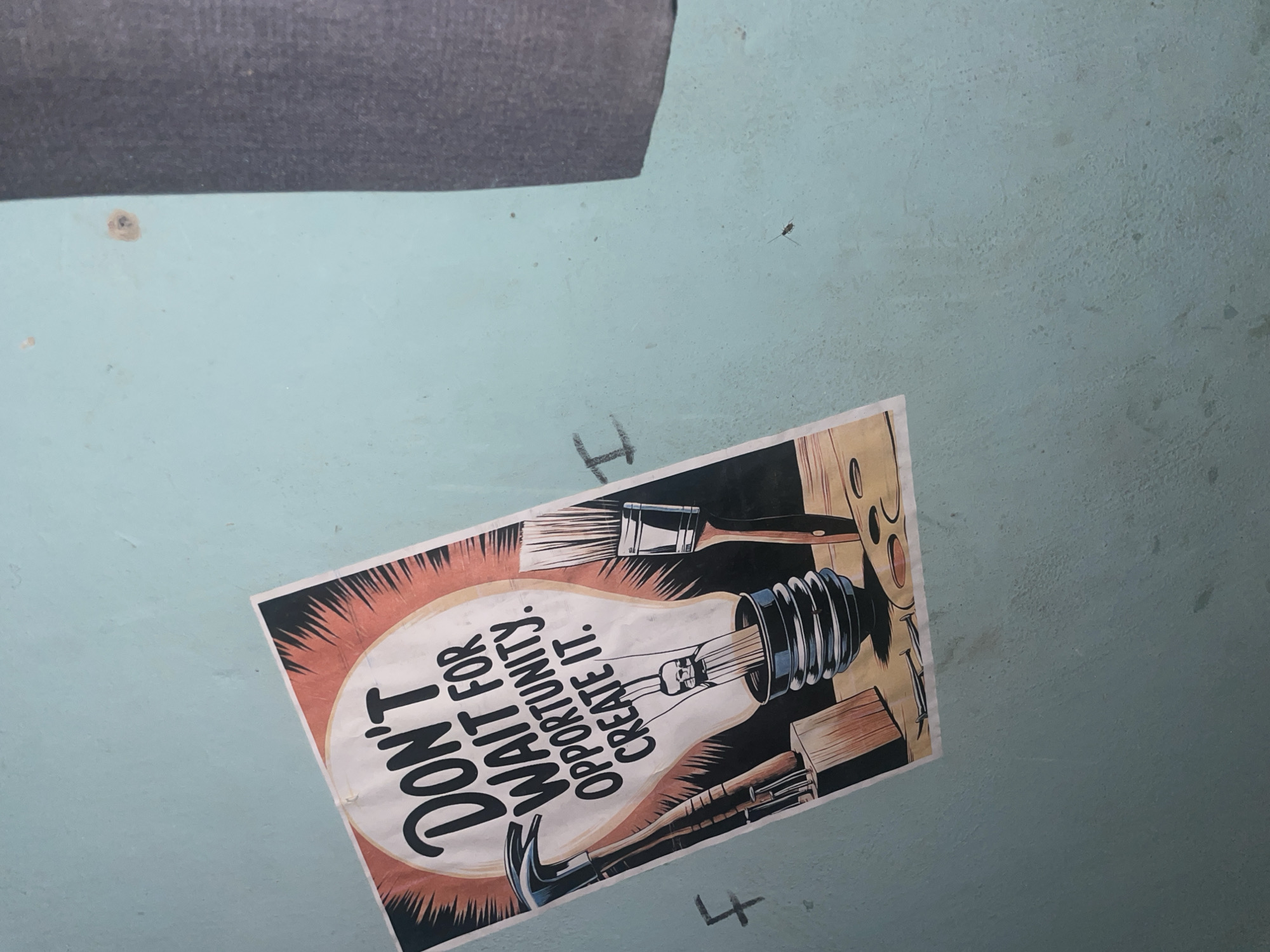 The image shows a poster on a light blue wall in Jalingo, Nigeria. The poster is tilted and features a graphic of a light bulb with the words "DON'T WAIT FOR OPPORTUNITY. CREATE IT." Tools like a hammer, paintbrushes, and nails are depicted around the light bulb, suggesting a theme of self-creation and hard work. The wall appears to be textured and has some marks and stains on it. A dark grey fabric is visible on the right side of the image, possibly a curtain or a piece of furniture. There are also some faint markings on the wall, including what appears to be a handwritten 'F' and 'I'. A small insect is visible on the wall near the top right.