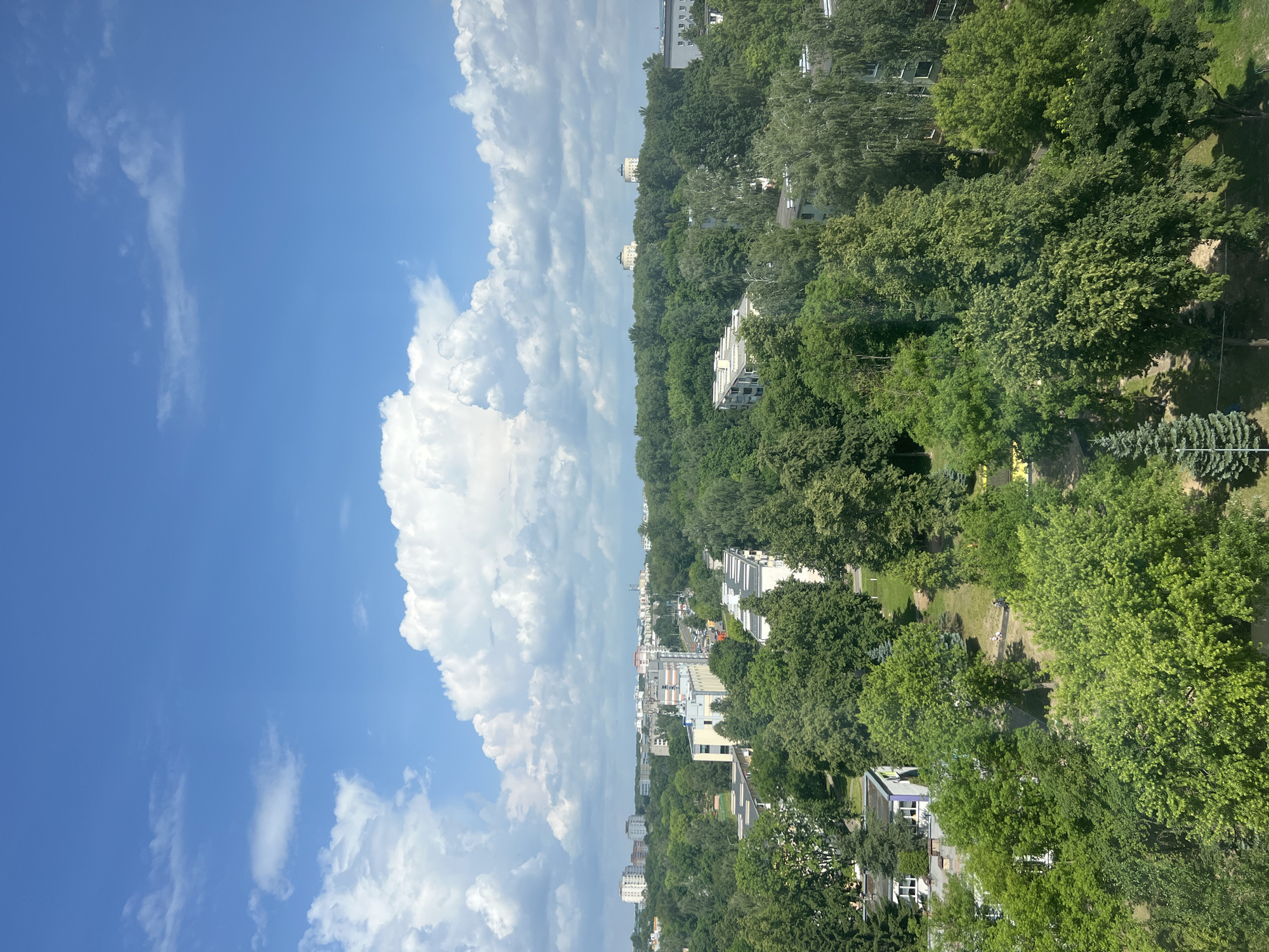 The image presents an aerial view of a city park on a sunny day with a bright blue sky dotted with white clouds. In the foreground, lush green trees dominate the landscape, casting shadows on the grass below. A paved path winds through the park, with two individuals walking along it. Further into the background, residential buildings and apartment complexes are interspersed with more trees, suggesting a mixed urban and green environment. In the distance, a skyline of larger buildings can be seen. The overall impression is one of a well-developed urban area with significant green spaces. The location is identified as Minsk, Belarus.