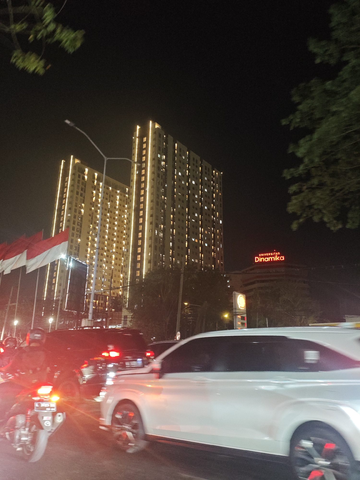 The image shows a nighttime street scene in Surabaya, Indonesia, likely near Universitas Dinamika. Two tall, brightly lit apartment buildings dominate the background, illuminated by vertical lines of warm yellow lights. In front of these buildings, on the left side, are several Indonesian flags, their red and white colors clearly visible. A street light with a curved pole is also visible between the buildings.
In the foreground, a traffic jam is evident. A white car is in the right lane, its wheels blurred due to motion. To the left of the white car, a dark-colored vehicle, possibly an SUV, is visible with its taillights illuminated. In the far left of the foreground, a motorcycle is present, also with its taillights and license plate illuminated. The motorcycle's license plate appears to read "L 3723 WNN". The scene is characterized by the glow of vehicle lights and the illuminated buildings, suggesting a busy urban environment at night. The presence of the university sign, "UNIVERSITAS Dinamika," in red neon lights on a building in the middle ground further confirms the location. Trees are visible on the right side of the image, partially obscuring the sky and the university building.