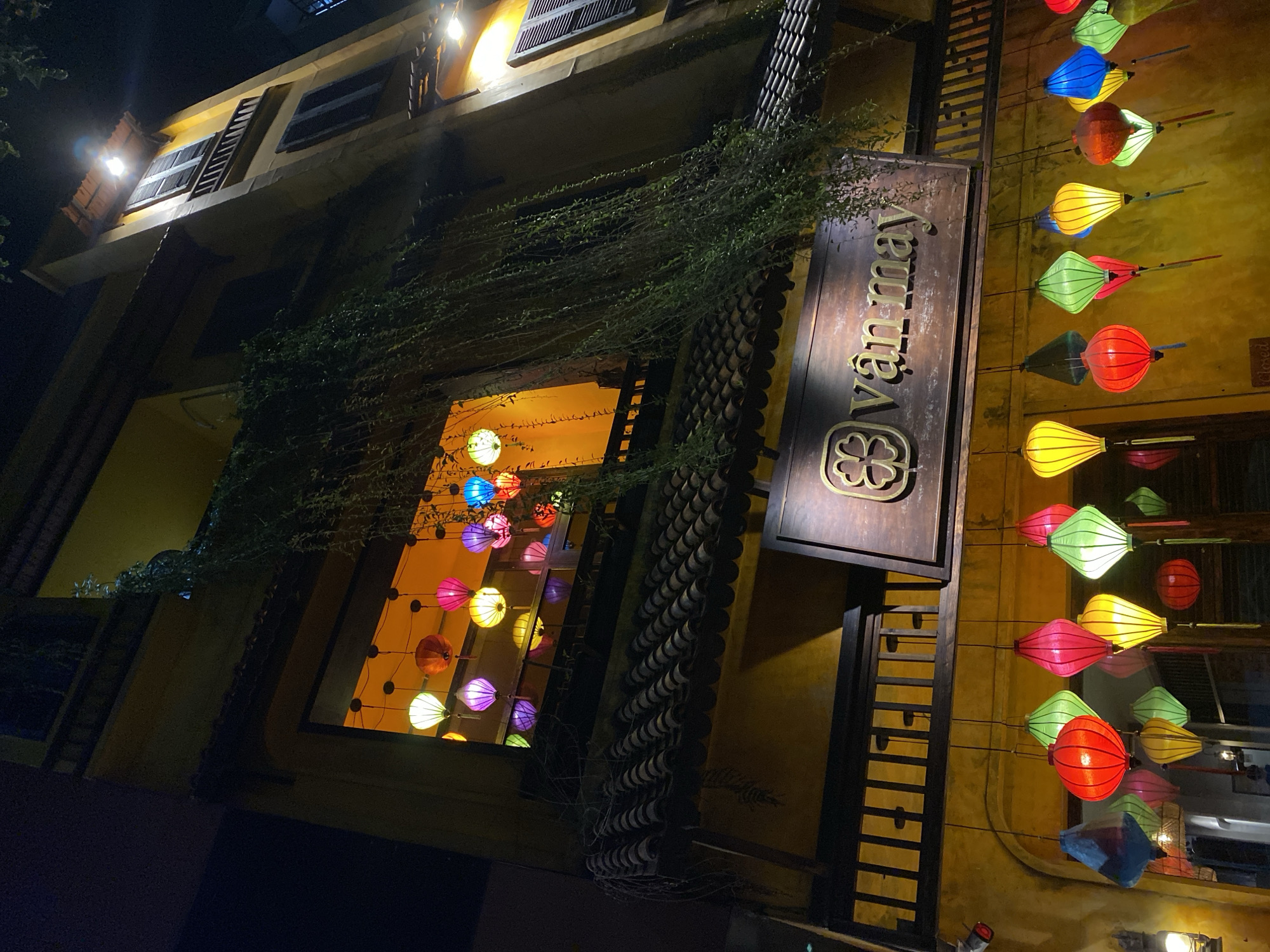The image shows the exterior of a building at night, likely in Đà Nẵng, Vietnam, decorated with numerous colorful lanterns. The building is yellow with traditional tiled roofs and has large windows illuminated from within, revealing more lanterns. Green vines cascade down the side of the building. A wooden sign with the text "vân may" is visible above a row of hanging lanterns in various shapes and colors, including red, green, yellow, pink, and blue. Some of the lanterns are diamond-shaped, while others are more rounded. The scene is dimly lit, with the lanterns providing the primary light source, creating a festive and inviting atmosphere. There are no people clearly visible in the frame. The architecture suggests a traditional Southeast Asian style, common in tourist areas.