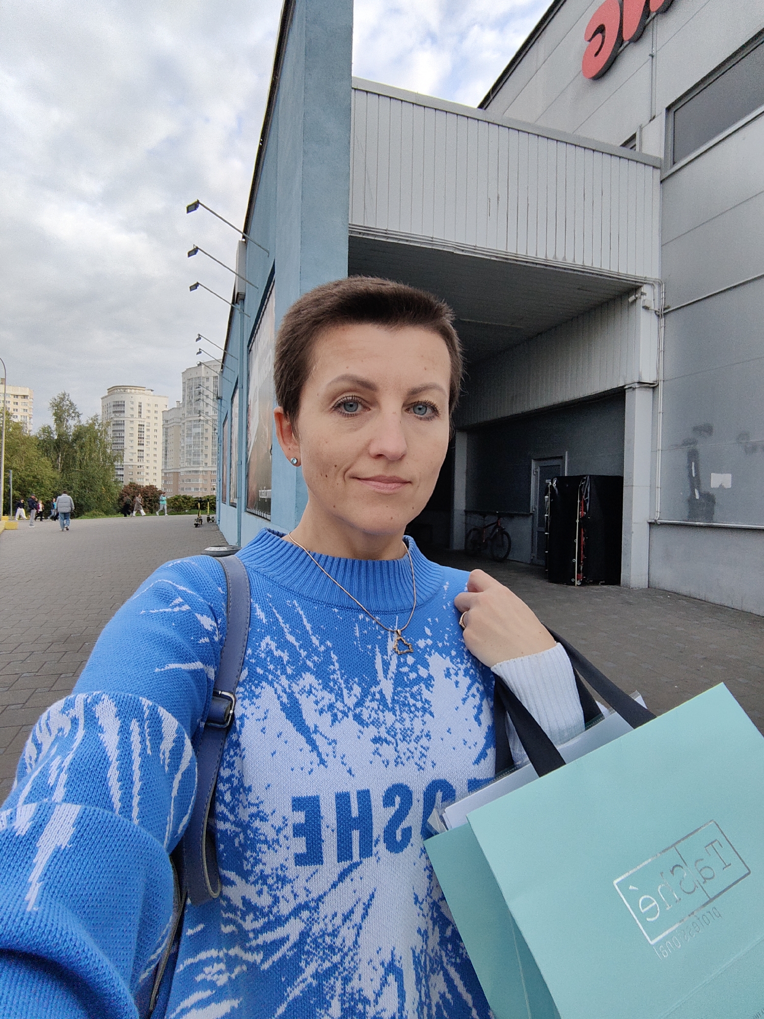 A young woman with short brown hair and blue eyes takes a selfie outdoors. She is wearing a blue and white patterned sweater, a silver necklace with a star pendant, and a silver ring on her right hand. She is holding two light blue shopping bags. The background shows a paved walkway, trees, apartment buildings, and the exterior of a building with a blue facade and grey metal siding. The logo on the shopping bag is in reverse. This scene is likely in the city of Minsk, Belarus, given the architectural style of the buildings and the presence of a shopping bag with a Cyrillic inscription.