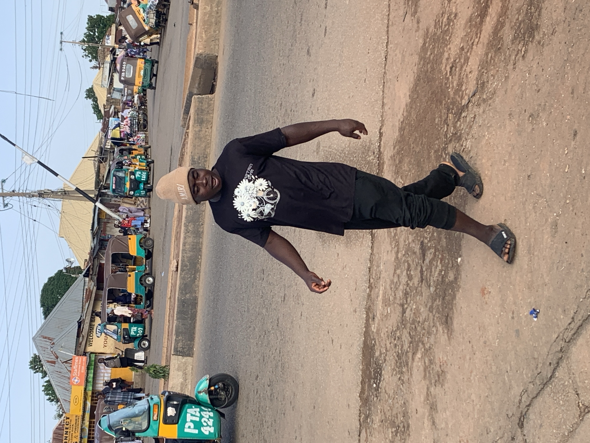 This media file captures a street scene in Gura Top, Nigeria. The primary subject is a young Black man wearing a black t-shirt with a white floral design and black pants, along with a beige beanie that has the word "AMIRI" visible. He is in a dynamic pose, leaning back with his right arm extended. The man is standing on a dusty, paved road, which is the setting for the image.
In the background, a busy street is depicted with various forms of transportation, predominantly auto-rickshaws, commonly known as "keke napep" or "marwa" in Nigeria. Several are parked or driving along the road, some displaying visible text such as "PTA 424", "PTA 307", and "PYA-7267". Buildings line the street, appearing to be shops or businesses, with some signage visible, including "StarTimes" and a "WELCOME BACK" banner. A few other people are scattered in the background, some near the vehicles and buildings.
The time of day appears to be late afternoon or early evening, suggested by the warm, diffused lighting and the shadows. The weather seems to be fair, with a clear sky visible above the buildings and power lines. The overall impression is one of everyday life in a bustling Nigerian town, with a focus on the man in the foreground and the vibrant street scene behind him. The man's expression suggests he might be posing or interacting with the person taking the photo.