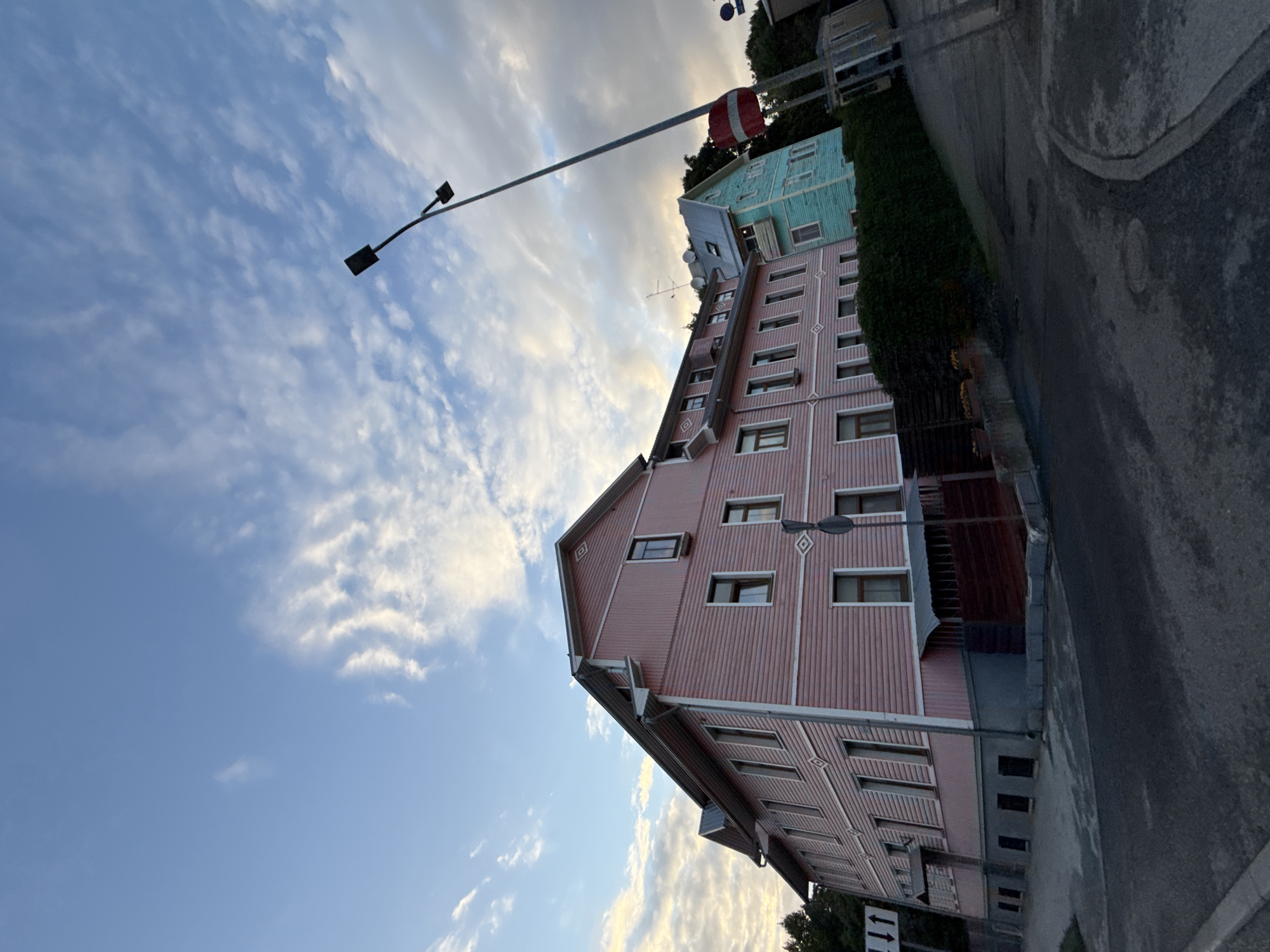 The image shows a street view in Tallinn, Estonia, with a distinctive pink wooden building on the left and a teal building behind it. The buildings appear to be old, multi-story residences or possibly businesses. A tall lamppost with two lights stands in front of the teal building, and a Latvian flag is visible attached to it. The street is paved with asphalt and curves to the right. There is a dense hedge separating the sidewalk from the buildings. The sky is partly cloudy with streaks of white clouds against a blue background, suggesting it is either early morning or late afternoon. There are no people visible in the image. The overall scene conveys a sense of quiet residential street in an older European city.