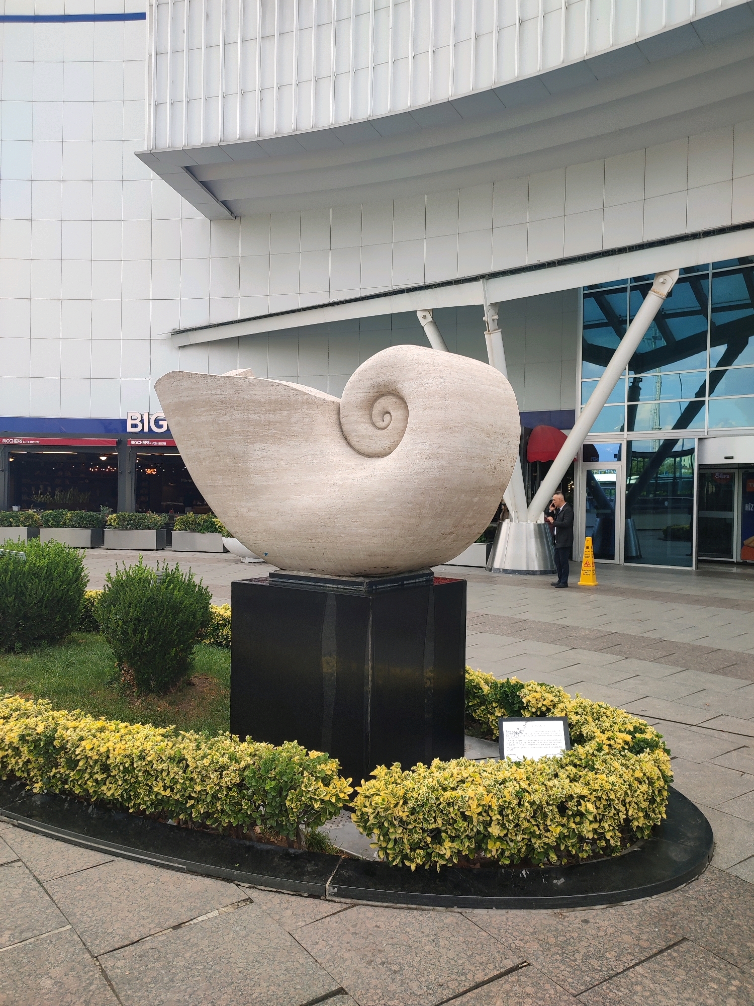 An outdoor, eye-level, full shot captures a large, light-colored, spiral sculpture resembling a nautilus shell. The sculpture is positioned on a dark rectangular base in front of a modern building with a glass facade and a white, paneled exterior. A man in a dark suit is standing to the right of the sculpture, holding a phone to his ear. He is standing next to a yellow cautionary cone. In front of the sculpture, there is a planter with manicured green and yellow bushes. The ground is paved with large, gray tiles. The building has a sign that reads "BIG" in blue and white letters. The scene appears to be in a public area, likely outside a commercial or office building. The overall impression is one of urban public art and architecture. This sculpture is located at the Nautilus Sculpture in Kadıköy, Turkey.
