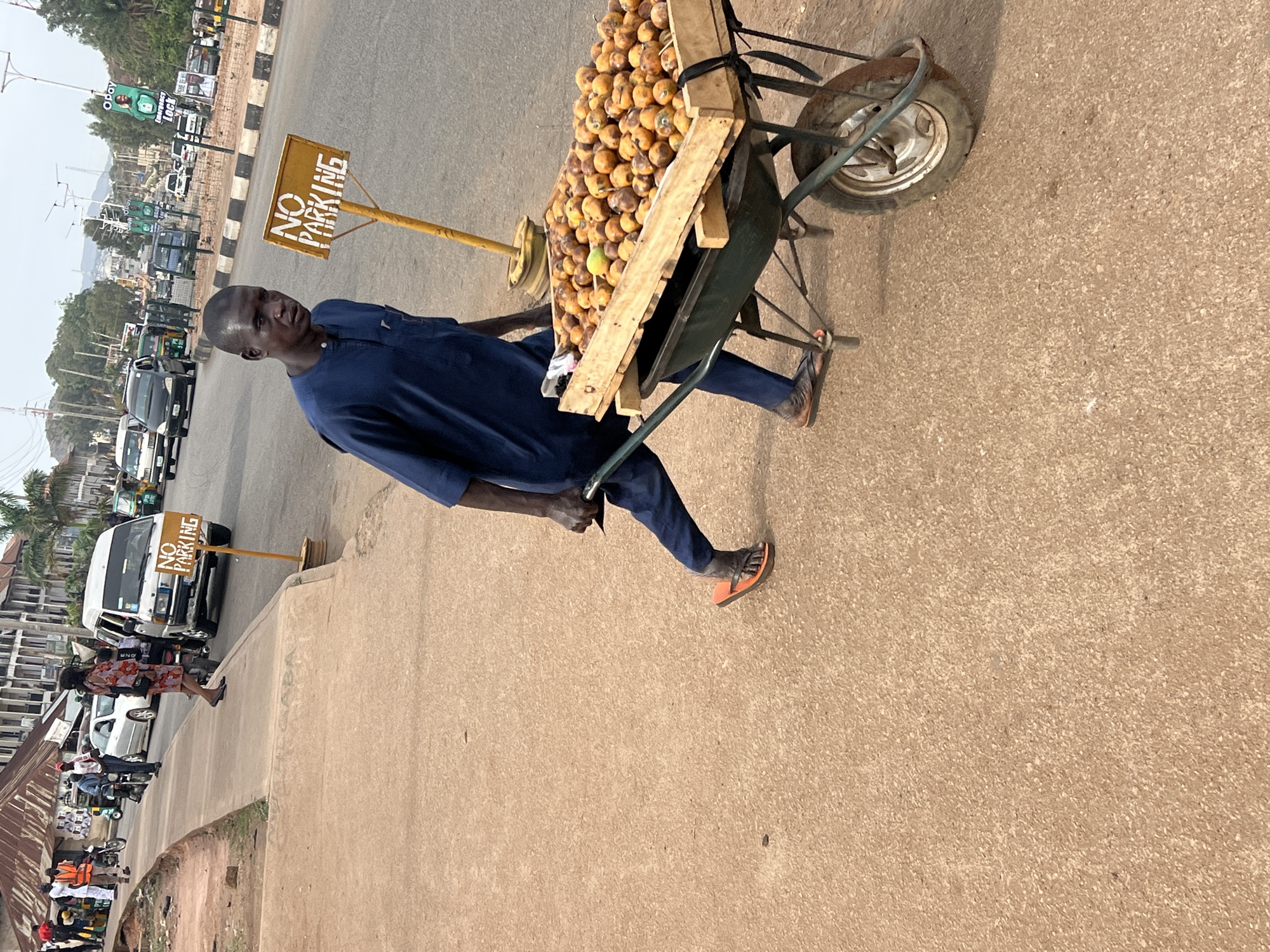 A photograph, rotated 90 degrees counter-clockwise, depicts a man pushing a wheelbarrow laden with fruit along a roadside in Jos, Nigeria. The central subject is a man dressed in a dark blue tunic and trousers, wearing orange flip-flops, actively pushing a green wheelbarrow. The wheelbarrow is modified with wooden extensions to hold a large mound of small, round, yellowish-orange fruits, likely local produce such as African star apples.
Prominently displayed to the right of the man is a yellow sign on a pole, bearing the text "NO PARKING". The background reveals a bustling urban environment under clear daylight conditions, with a bright, slightly hazy sky. Various vehicles, including cars and vans, are visible on the road and parked along the side, suggesting traffic and activity. Buildings and other structures line the street in the distance. A green billboard in the background partially shows the text "OPAY" and "Lock". The scene captures a moment of street vending or transportation of goods in a vibrant city setting.