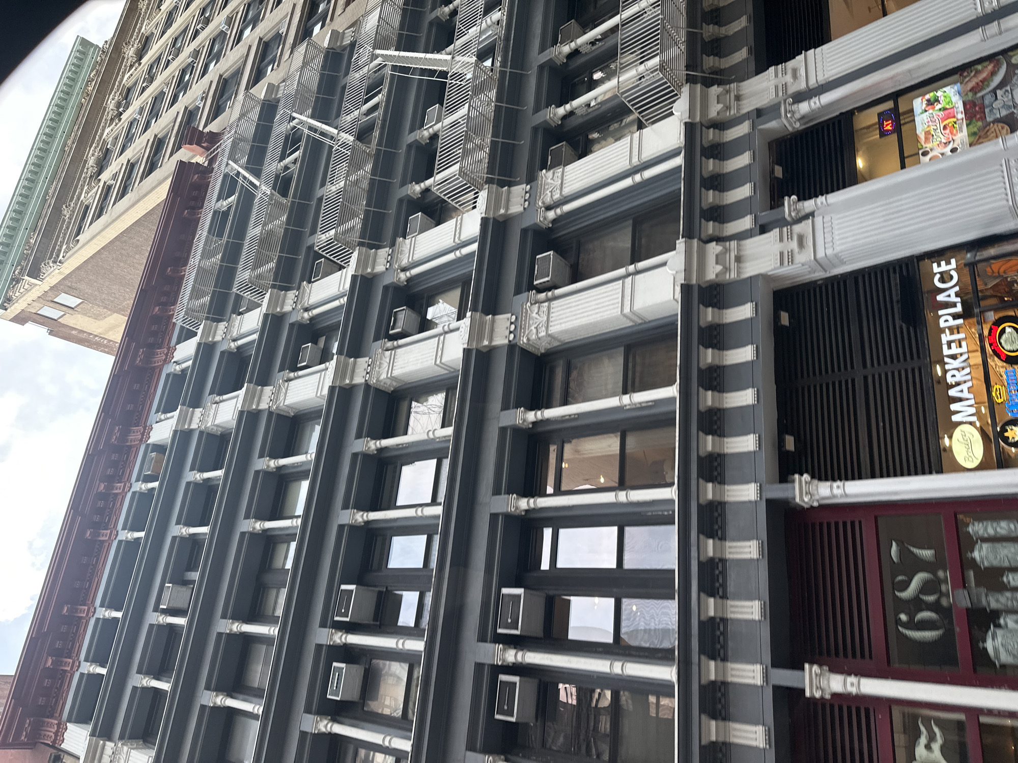 This is a ground-level, eye-level, wide-angle shot of the facade of a multi-story building in New York City, United States of America. The building has a dark grey exterior with white decorative columns between windows on the upper floors. Several windows are visible, many with air conditioning units protruding from them. The building also features fire escapes on some of the upper floors. The ground floor of the building has storefronts. One storefront has the number "687" displayed prominently on a dark red background. Another storefront displays signage for "MARKET PLACE" and features a display of food items. The architectural style suggests it is an older building, possibly from the late 19th or early 20th century, common in areas like SoHo or the Lower East Side. The sky is visible at the top of the image, suggesting it is daytime. There are no people clearly visible in the image. The overall scene is one of urban commercial architecture.
