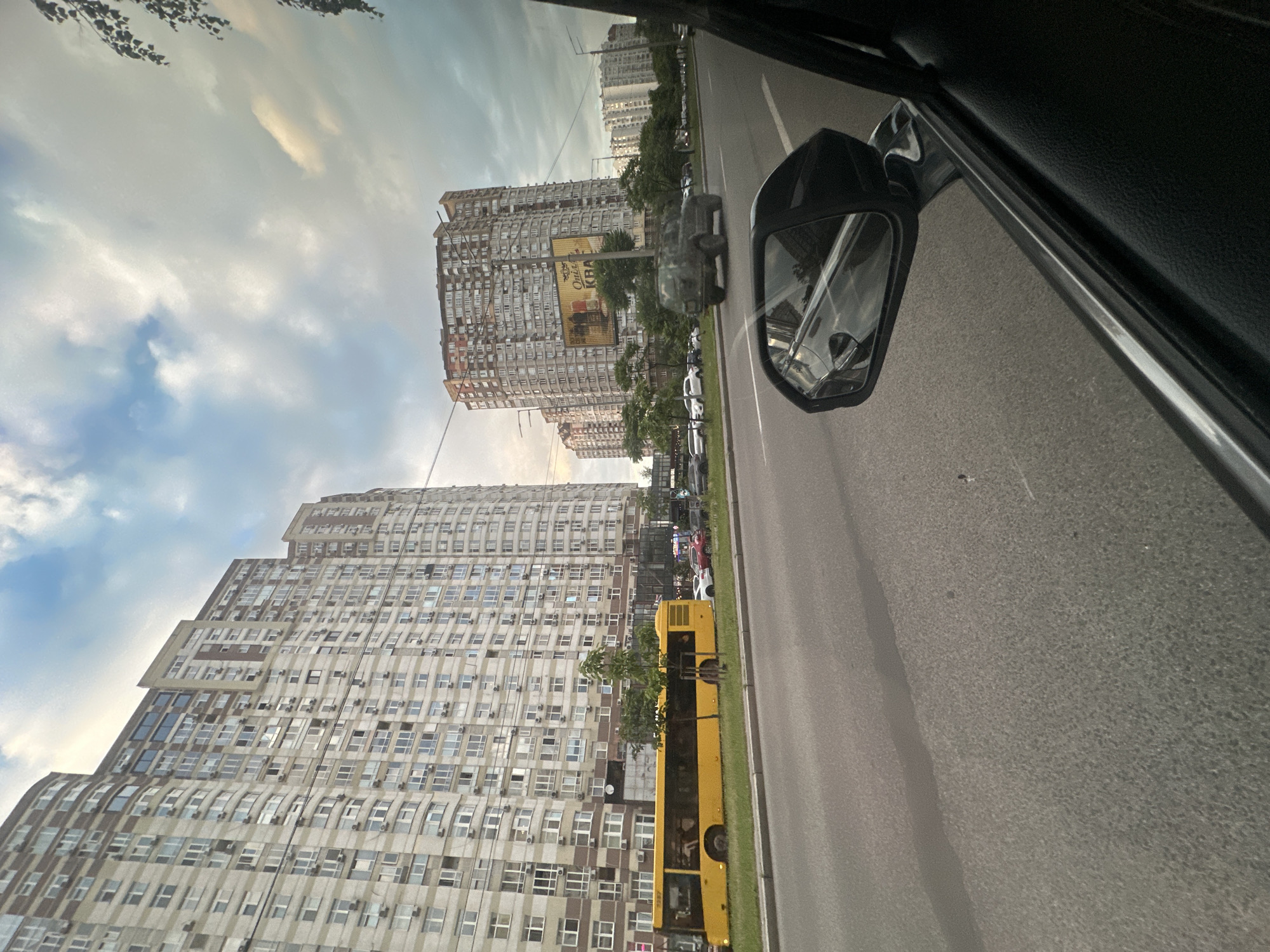 This media file captures a view from a car in Kyiv, Ukraine. The scene is an urban street with tall, imposing apartment buildings lining one side of the road. The buildings are distinct in their architectural style, with many windows and balconies, giving them a textured appearance. A bright yellow bus is parked on the side of the road, partially obscured by trees. Other cars are visible on the road and parked along the sidewalk, suggesting a typical day in the city.
The sky above is filled with clouds, with patches of blue sky visible. The lighting suggests it is either early morning or late afternoon, with a soft, diffused light. The overall mood is one of everyday urban life.
A car's side mirror is prominently visible on the right side of the frame, reflecting the sky and the street. Above the apartment buildings, some trees with dark leaves are seen against the sky, possibly from the perspective of another vehicle or a building.
There is visible text on a large billboard attached to one of the apartment buildings. The text appears to be in Cyrillic script, and parts of it are legible, including "Onl" and "KVAA". The context of this billboard is unclear, but it adds a layer of local detail to the scene. The image is oriented vertically, with the top of the frame showing the sky and the bottom showing the road and buildings.