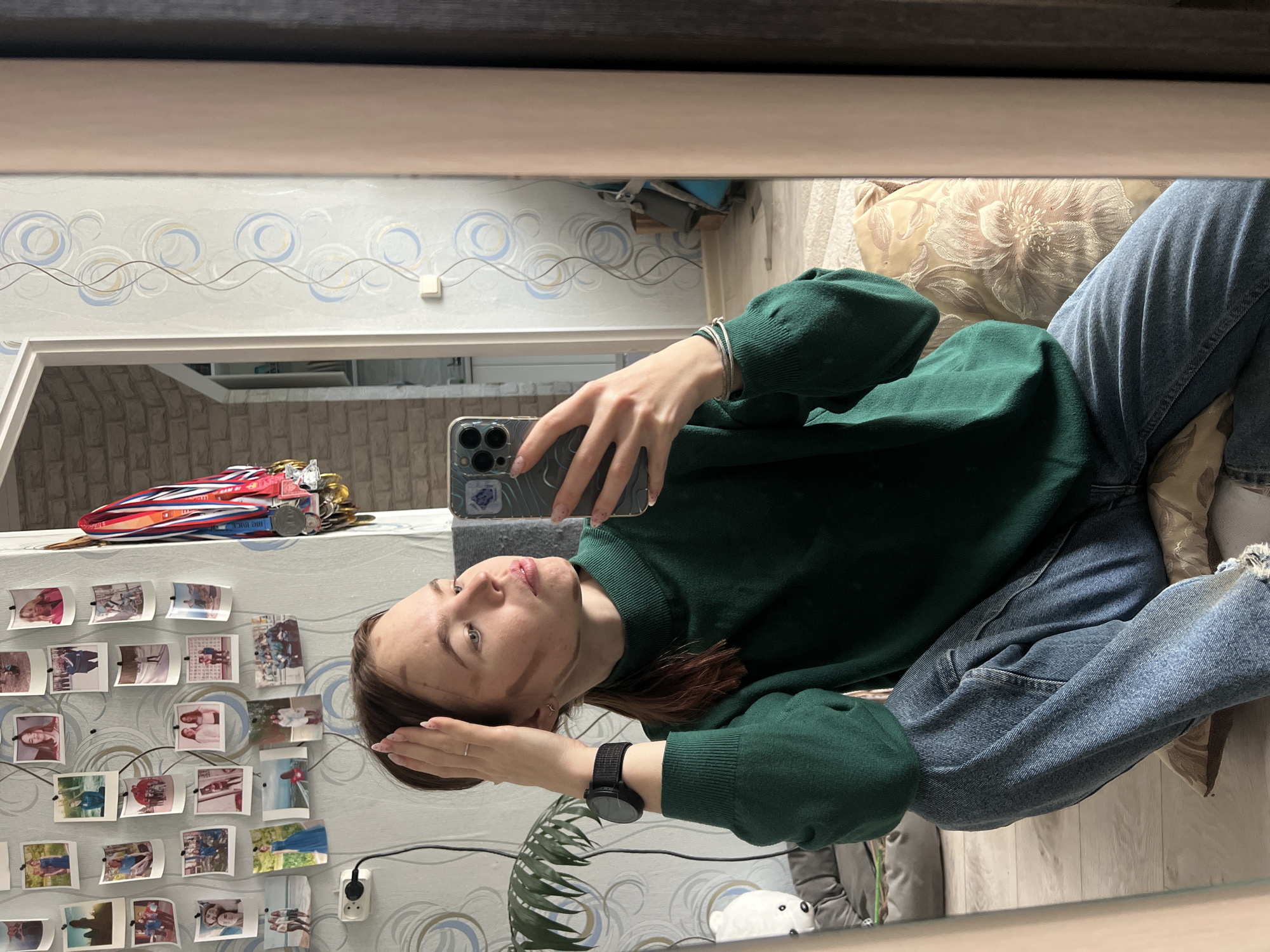 A young woman is taking a selfie in a mirror. She is wearing a green sweater, blue jeans, and a black smartwatch. The mirror reflects a room with wallpaper featuring a blue and gold swirl pattern. There are also pictures attached to the wall. A shelf is visible with a collection of medals on top. In the foreground, near the bottom of the mirror's reflection, a plant and a white figurine are noticeable. The location is likely Chaikovsky, Russia, based on the subject's social media profile.