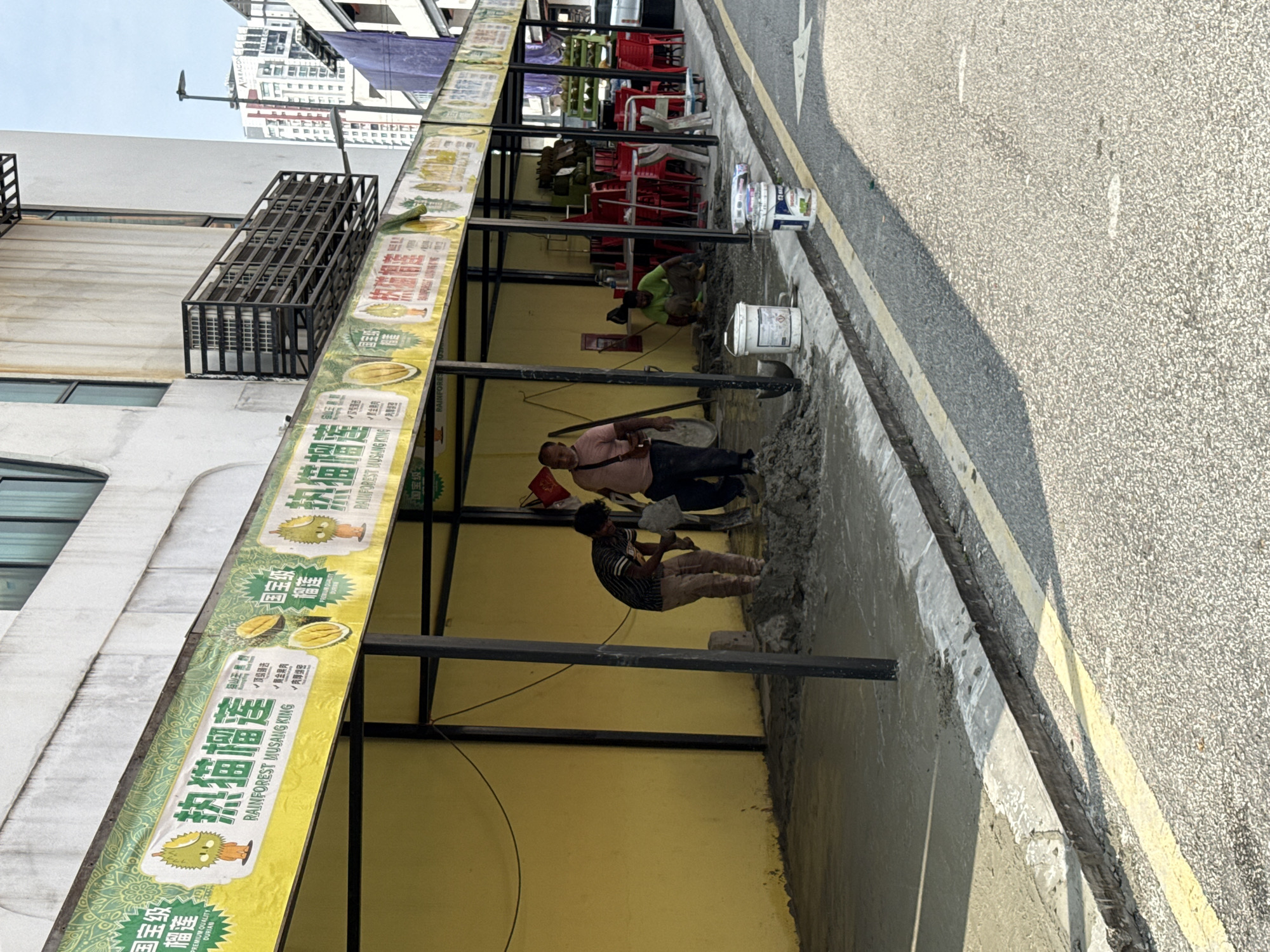 Here is a description of the image:
The scene depicts an outdoor setting that is likely a commercial area in Kuala Lumpur, Malaysia, as suggested by the signs advertising "Rainforest Musang King Durian" in Chinese characters. A yellow-painted building is adorned with promotional signs, showcasing durian fruits. Several individuals are engaged in construction or repair work along the sidewalk, near the road. The area appears to be under renovation, with concrete or other materials scattered about. The road is visible, marked with yellow lines. In the background, chairs are visible under the building's structure and a building is visible in the distance.