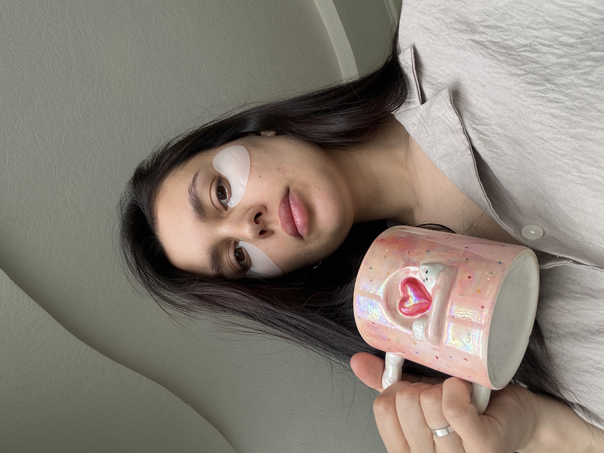 A young woman is captured in a close-up, head-and-shoulders portrait, lying or reclining indoors, likely in Chaikovsky, Russia. She has long, dark hair and a natural complexion, with light-colored under-eye patches visible below both eyes, suggesting a skincare routine. Her lips have a soft, natural pink tint. She wears a light grey or beige collared shirt, possibly made of a shimmering, silk-like fabric.
She holds a distinctive pink mug with an iridescent, pearlescent finish, adorned with small colorful confetti-like dots. The mug features a raised 3D design of a white, cartoon-like figure, possibly a cat or rabbit, hugging a prominent, glossy red heart. Her hand, visible holding the mug, shows a simple silver ring on her left ring finger. The background is a plain, light grey wall. The lighting appears soft and natural, suggesting daytime. The overall impression is one of relaxation and self-care. No visible text is present in the image.