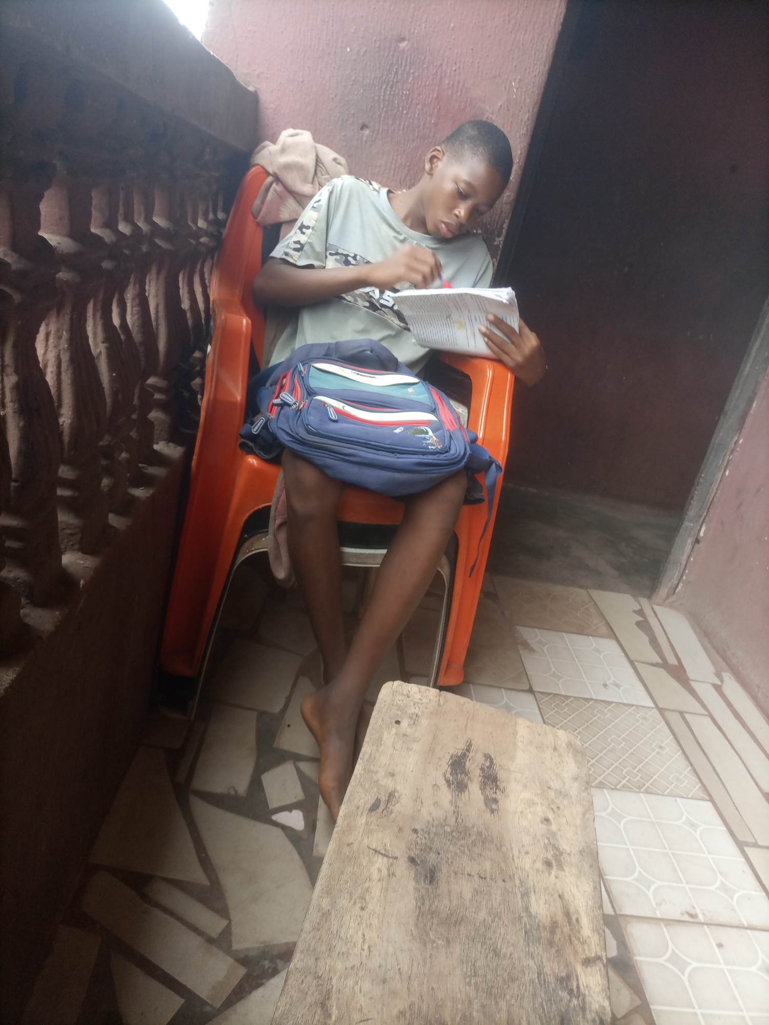 A young Black male sits on an orange plastic chair, holding a book and a pen, appearing to be studying. He wears a grey t-shirt with a camouflage pattern on the sleeves. A blue and red backpack rests on the seat of the chair behind him. The chair is positioned on a tiled floor, partially covered by a rough wooden plank. Behind the chair, a textured reddish-brown wall is visible, with a dark doorway or opening to the right. To the left, a decorative, carved stone balustrade runs along a wall. The lighting suggests an outdoor or semi-outdoor setting, possibly a balcony or porch.
