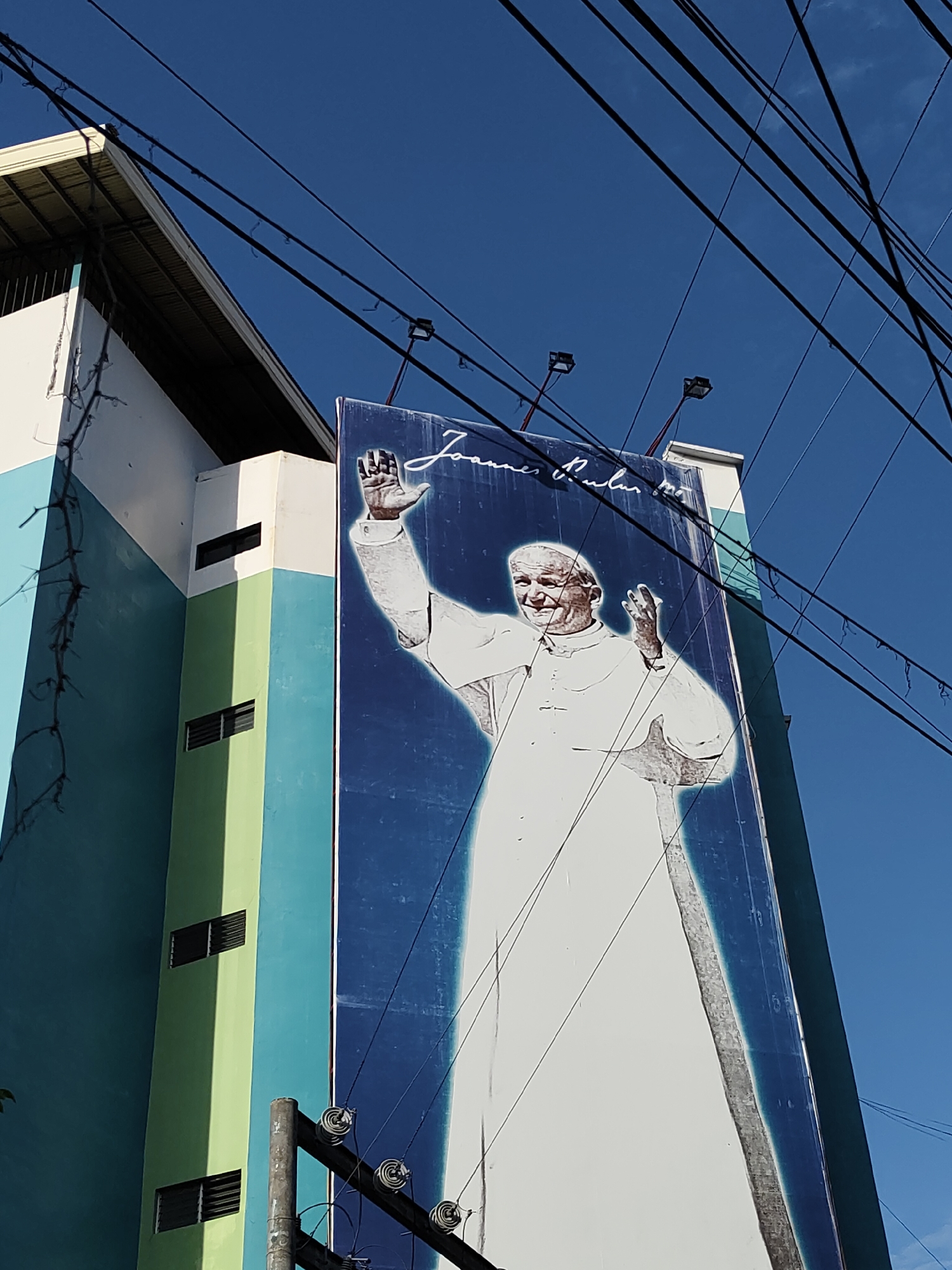 A large outdoor billboard prominently displays a smiling depiction of Pope John Paul II, clad in a white papal cassock with both hands raised in a welcoming or blessing gesture. The billboard features a blue background with a radiant white glow outlining the Pope's figure. Above his image, the cursive text "Joannes Paulus II" is clearly visible.
The billboard is affixed to or stands next to a multi-story building, which is painted in alternating vertical stripes of light blue, white, and light green. Several small, rectangular windows with horizontal louvers can be seen on the building's facade. The scene is set under a clear, vibrant blue sky, suggesting bright daytime conditions, likely morning or early afternoon due to the strong sunlight. Numerous dark power or communication lines crisscross the upper part of the frame, spanning across the sky and above the billboard. Two floodlights are mounted on the top edge of the billboard structure, presumably for illumination after dark. A utility pole with visible insulators is partially visible in the bottom right corner of the frame. This public display of a revered religious figure is situated in what appears to be an urban or semi-urban environment, consistent with Davao City, Philippines.