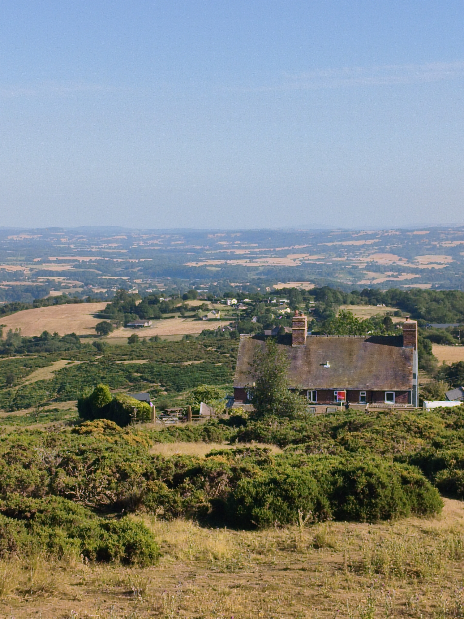 The image depicts a panoramic view from an elevated position in Cleehill, United Kingdom, showcasing a rural landscape under a clear blue sky. In the mid-ground, a prominent brick house with a tiled roof and two chimneys stands on a hillside covered in dense green vegetation. The house has several windows, and a red sign is visible in one of them. The surrounding terrain is characterized by rolling hills with patches of dry grass, fields of various shades of brown and green, and clusters of trees. In the background, a vast expanse of countryside stretches to the horizon, featuring more undulating hills and agricultural fields. The overall scene conveys a sense of tranquility and rural serenity. There are no people or animals visible in the image.