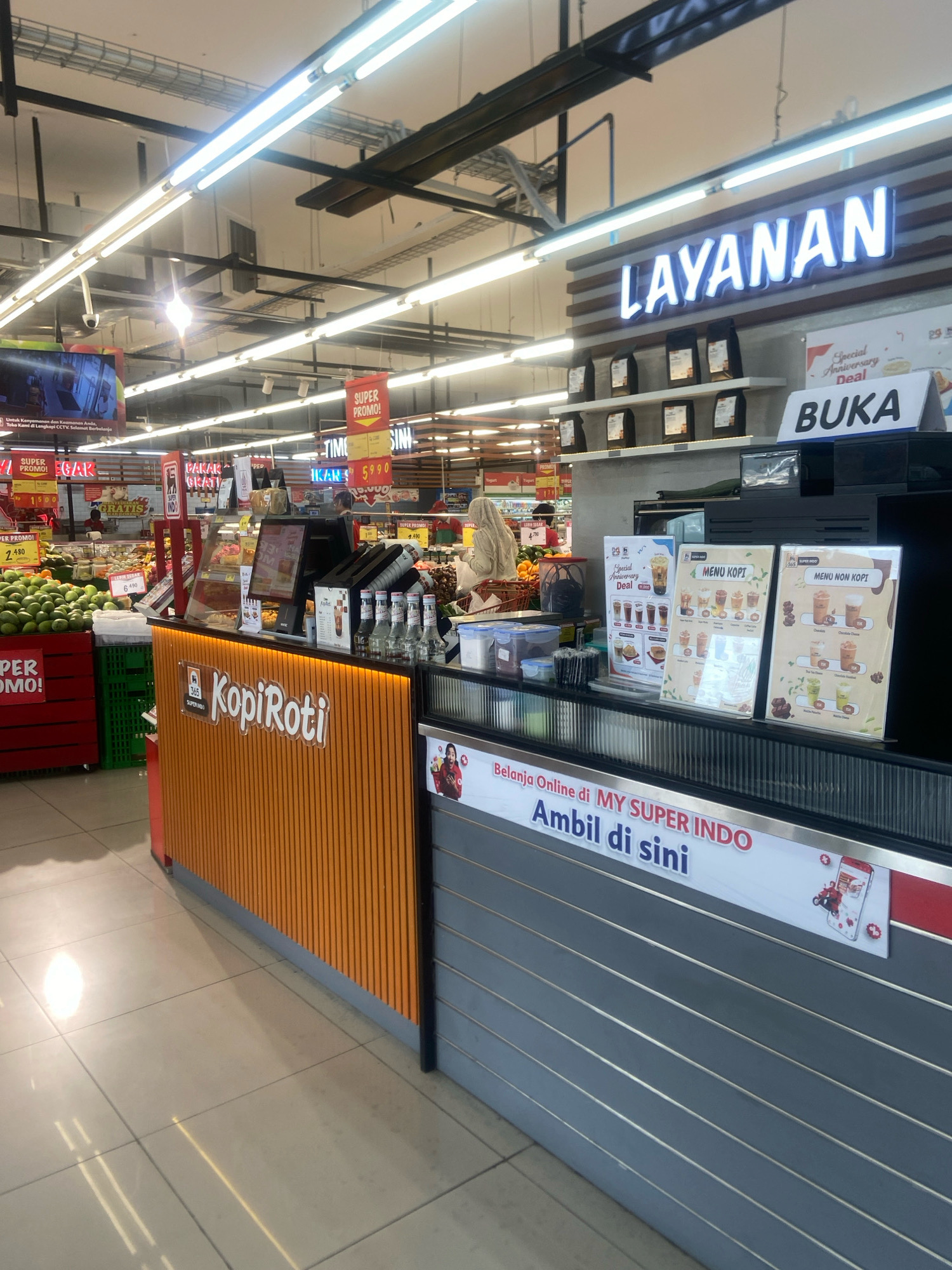 The image shows an interior scene of a supermarket in Ngaglik, Indonesia. The foreground is dominated by a service counter with an orange textured front and a grey ribbed facade. On the counter, there are several bottles of drinks, a cash register, and menu boards displaying coffee and non-coffee beverages. A prominent sign above the counter reads "LAYANAN" in large, illuminated blue letters. To the right of the counter, a sign indicates "BUKA" in bold lettering.
The middle ground of the image reveals shelves stocked with packaged goods, likely coffee bags, behind the counter. Further back, aisles of the supermarket are visible, with shelves filled with various products. There are also signs indicating promotions like "SUPER PROMO!" and product names with prices. Fruit, specifically avocados, is displayed in red and green crates on the left side of the image. A person wearing a headscarf is partially visible behind the counter, attending to customers or duties.
The environment is brightly lit by fluorescent lights on the ceiling. The floor is tiled and reflects the lighting. A sign on the grey facade of the counter reads "Belanja Online di MY SUPER INDO Ambil di sini," indicating an online shopping pickup point. The overall impression is a well-organized and active retail environment.