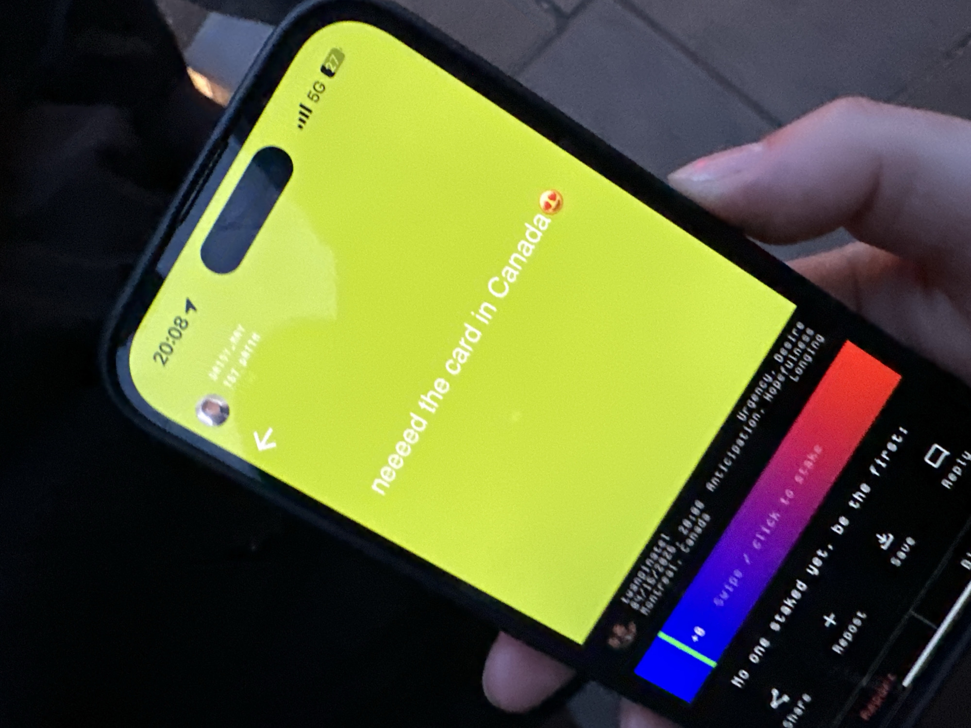 The image captures a close-up view of a person's hand holding a modern smartphone, likely an iPhone given the distinctive Dynamic Island cutout. The screen is brightly lit, contrasting with the dark, out-of-focus background, suggesting a low-light setting. The phone's status bar shows the time as "20:08" (8:08 PM), a 5G network connection, and 27% battery life.
The primary content on the phone screen is displayed against a vibrant yellow background, featuring the text: "neeeed the card in Canada" accompanied by a heart-eyes emoji, indicating a strong desire. Below this, faint text mentions a user "tuanpinatel," a timestamp "84/16/2626, 20:08" (the year likely a typo), and "Montreal, Canada." Adjacent to this information, a list of emotions is displayed: "Urgency, Desire, Anticipation, Hopefulness, Longing."
Further down the screen, a colorful gradient bar (transitioning from blue to purple to red) prompts "Swipe / click to stake," with a value of "-.8" indicating some sort of progress or score. Below this, the phrase "No one staked yet, be the first:" encourages user engagement. At the very bottom, common social media interaction options are visible: "Share," "Repost," "Save," and "Reply."
The image depicts an individual engaging with a mobile application, expressing a clear longing for a product or service to be introduced in Canada. While the content on the phone references Montreal, Canada, the image's provided location context is London, United Kingdom.