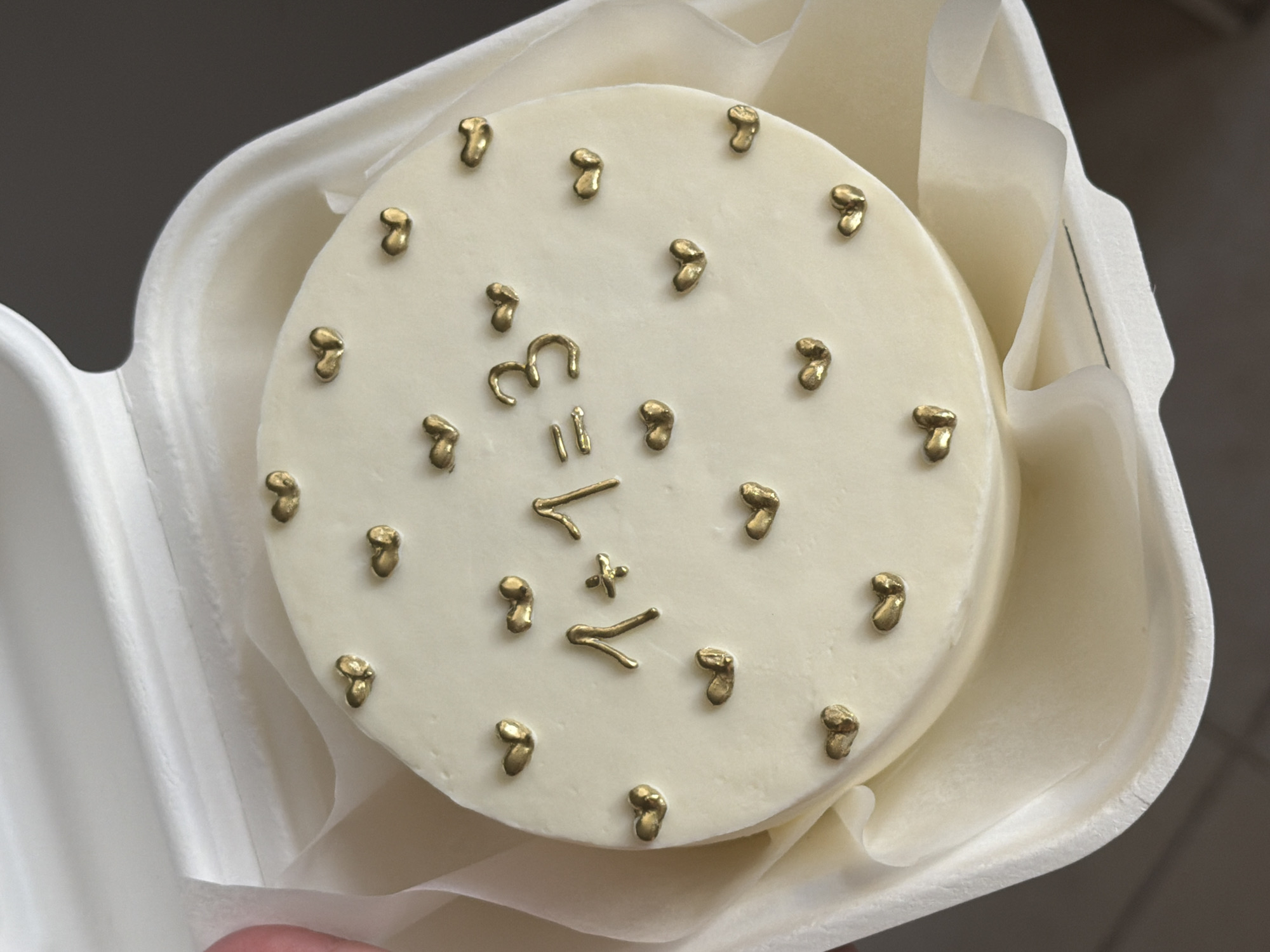 A top-down, slightly angled view captures a small, round white cake presented in a white, molded fiber container. The cake's smooth, pristine surface is adorned with numerous small, irregularly shaped, gold-colored decorations.
Prominently featured in the center of the cake is a mathematical equation formed by these gold pieces: "3 = 1 + 2". The numbers '3', '1', and '2', along with the equals sign and plus sign, are clearly rendered in the metallic gold, standing out against the white frosting. Surrounding this central equation, additional gold pieces, resembling small nuggets or abstract shapes, are scattered decoratively.
The cake rests on a layer of crinkled, translucent parchment or tissue paper that lines the inside of the container, providing both cushioning and an elegant presentation. The container itself appears to be made from a biodegradable material. A portion of a person's thumb is visible in the bottom left, holding the container, suggesting the cake is being handled or has just been received.
The background is an indistinct, blurry dark greyish-brown, indicating an indoor setting. The lighting is bright and even, casting subtle shadows that highlight the textures of the cake, decorations, and packaging. The overall aesthetic is clean and modern. The location context provided for this media is Tel-Aviv, Israel.