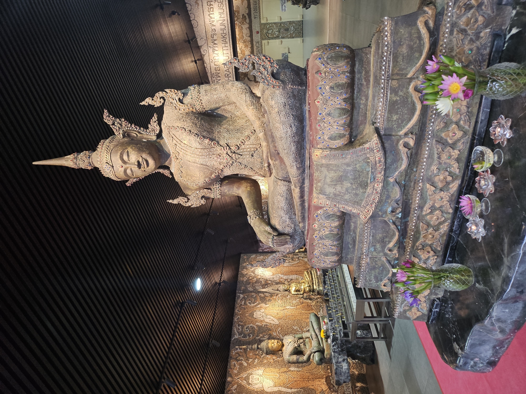 In the city of Na Chom Thian, Thailand, an indoor scene features a large, ornate statue of Buddha seated in a meditative pose. The Buddha statue is made of metal, with intricate carvings and colorful embellishments. In the foreground, a vase of lotus flowers and a few candle holders are placed on a dark marble surface. Behind the main Buddha, smaller Buddha statues are visible, as well as a mosaic wall depicting figures. The ceiling of the room is made of dark wood slats. The overall atmosphere suggests a temple or a museum dedicated to Buddhist art.