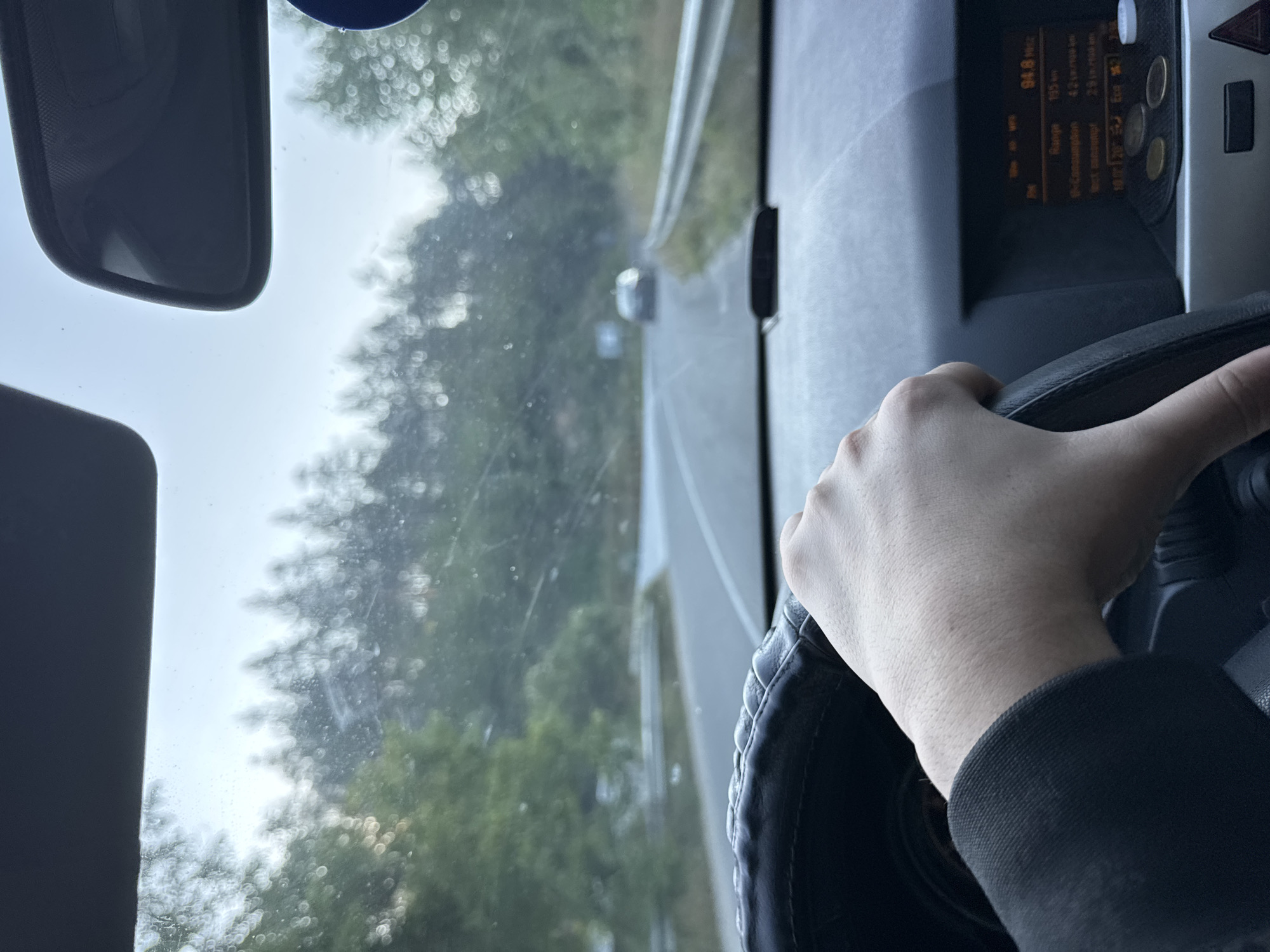 The image shows a first-person perspective from the driver's seat of a car on a road. The driver's hand is visible gripping the steering wheel. The dashboard of the car is in the foreground, with a display screen showing "94.8 MHz" and other text. The road ahead is visible, with trees lining both sides. A silver car is in the distance on the road. The sky is overcast. The location is identified as the city of Taran, Bulgaria.