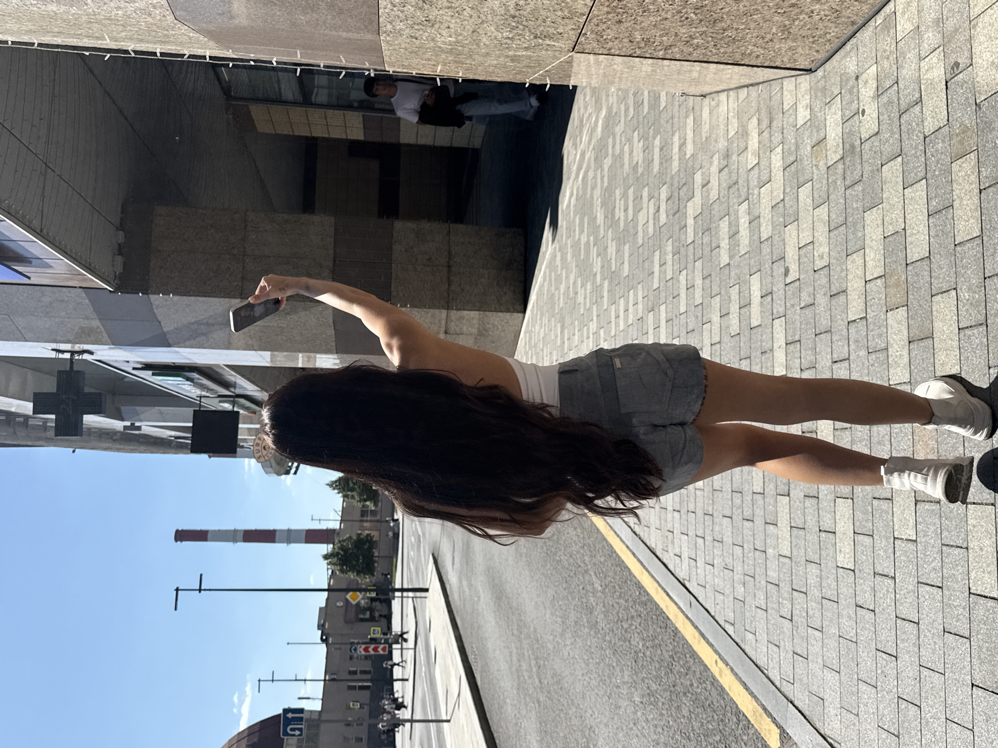 A young woman with long, dark hair is walking on a pedestrian walkway in Moscow, Russia, holding up her smartphone as if to take a picture or record a video. She is wearing a white top and grey shorts, with white ankle boots. The walkway is paved with rectangular grey and beige stones and runs alongside a street with a yellow line marking the curb. In the background, there is a multi-story building with a stone facade and a large glass window. Across the street, a red and white striped chimney stack is visible against a clear blue sky. Other pedestrians and buildings are also visible in the distance. To the right of the woman, a man in a white t-shirt and light-colored pants is walking away from the camera. The scene appears to be a sunny day in an urban environment.