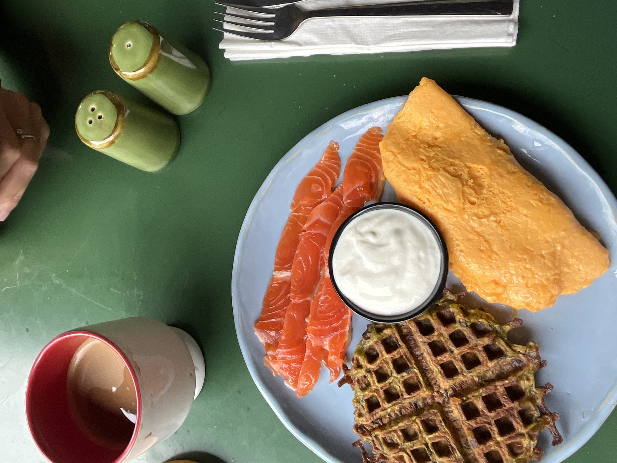A high-angle shot shows a breakfast setting on a green table. In the foreground, a light blue plate holds a meal consisting of a savory waffle, thinly sliced smoked salmon, a dollop of white cream, and a folded omelet. To the right of the plate, a white napkin is topped with a fork and knife. Further back, a white mug with a red rim contains a light brown beverage, possibly coffee or tea. Two green salt and pepper shakers with brown rims sit to the right of the mug. A person's hand, with a silver ring on one finger, is visible in the upper right corner of the image, reaching towards the shakers. The environment is a restaurant or cafe in Mengwi, Indonesia.
