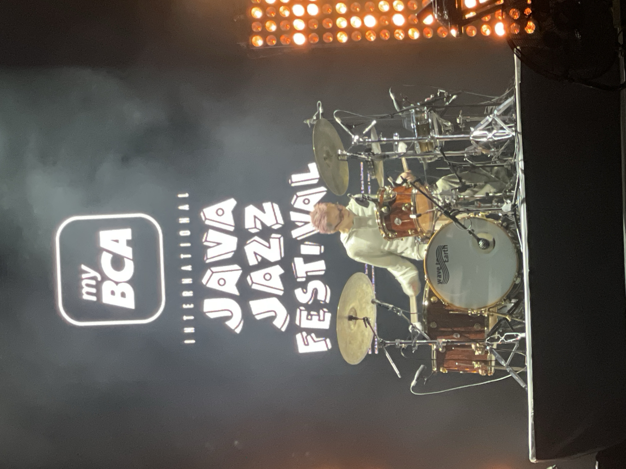 The image captures a performance at the International Java Jazz Festival, likely in Salembaran, Indonesia. The backdrop displays the festival's logo featuring "my BCA" and "JAVA JAZZ FESTIVAL INTERNATIONAL" in large, white lettering against a black background. A drummer is positioned behind a complex drum set with multiple cymbals and toms. The drum set includes a bass drum with "wave Earth" logo on its head. The lighting above the stage consists of numerous circular lights. The overall environment suggests a concert setting.