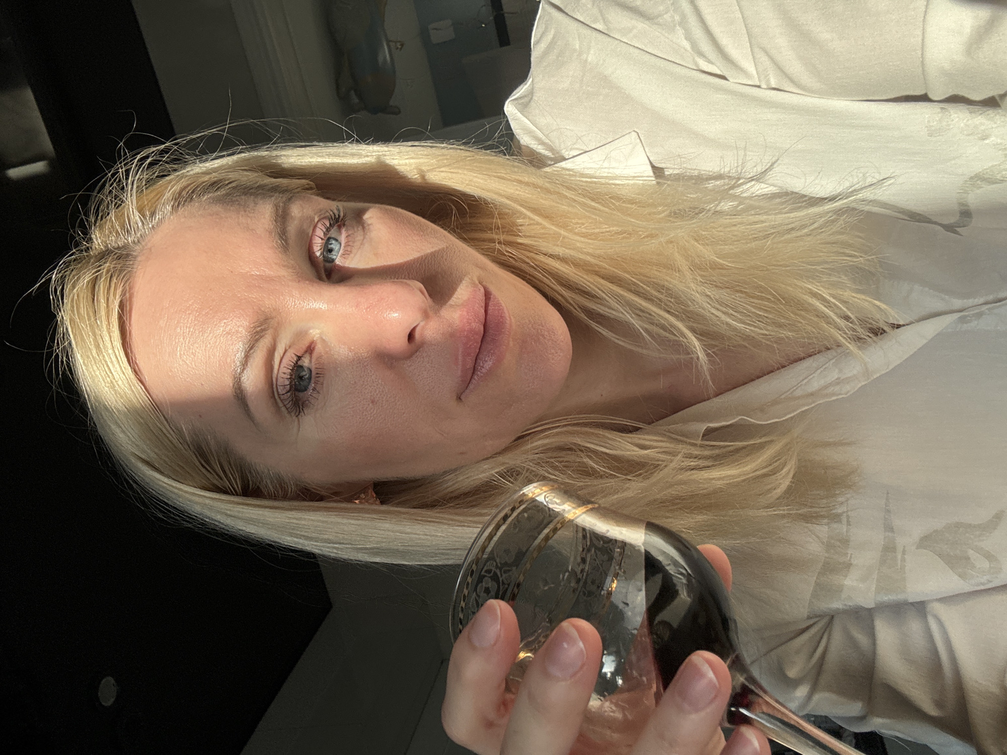 A close-up portrait shows a woman with blonde hair and light blue eyes, holding a wine glass. Her face is angled slightly, with direct natural light, likely sunlight, illuminating her from the left, creating distinct shadows on her forehead and nose. Her expression appears calm and neutral, with her gaze directed softly towards the viewer. Her lips are slightly parted.
She holds a decorative wine glass, containing a dark red liquid, in her right hand at the bottom right of the frame. The glass features an intricate pattern near its rim. She is wearing a light-colored, possibly off-white, shirt or blouse, visible on the right side and extending behind her head, characterized by folds and subtle patterns.
The background on the far left is dark, suggesting an indoor setting with dark elements. On the right, behind the light fabric, a blurred reflection or indistinct room details are faintly visible, also reflected in the glass. The strong lighting indicates it is daytime. No visible text is present in the image. The location is unknown, in Russia.