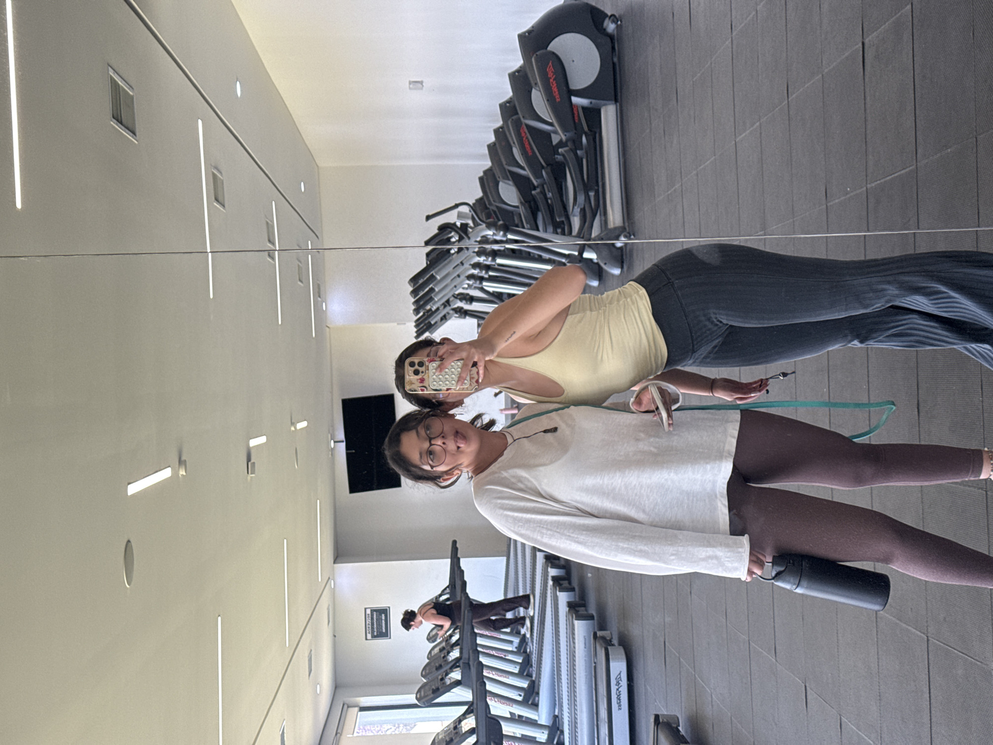 The image depicts a mirror reflection of two women in what appears to be a gym or fitness center. The woman closest to the foreground is wearing glasses, a long-sleeved white shirt, and brown leggings, and she is holding a black water bottle. She is looking at the camera and sticking out her tongue. Behind her and slightly to the left, the second woman is holding a phone with a patterned case, presumably taking the photo. She is wearing a yellow tank top and black pants. In the background, there are rows of treadmills and elliptical machines, some of which are in use by other people. The environment is brightly lit, with modern architecture including linear wall lighting and a dark rectangular screen or TV. Based on the location provided (San Andrés Cholula, Mexico), it is possible that the gym is a part of a fitness center or health club within the city.