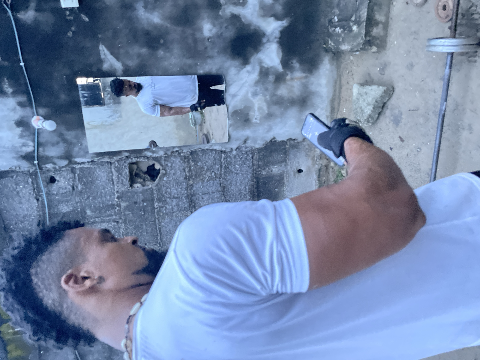 In Ado, Nigeria, an individual with a distinctive mohawk hairstyle stands outdoors in what appears to be a makeshift gym area. The person, wearing a white t-shirt, black gloves, and black pants visible in a reflection, is looking down at a smartphone held in their left hand. A barbell with weights is positioned on the ground in the foreground. The background features a rough, textured wall and a mirror reflecting the individual. The environment suggests an urban or semi-urban setting with elements of disrepair.