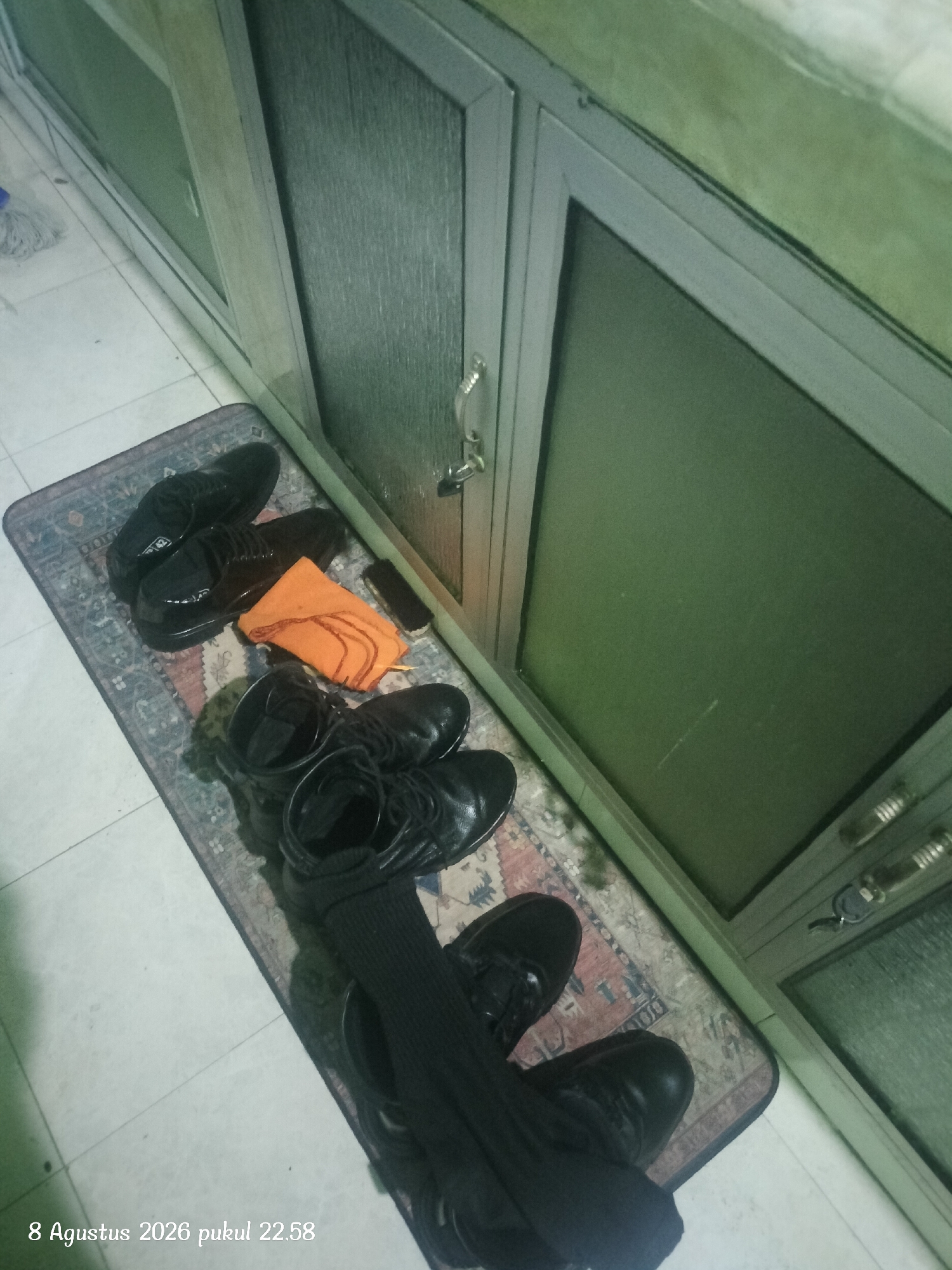 A still, high-angle, medium shot captures several pairs of black dress shoes arranged on a patterned rug placed on a tiled floor. The rug is positioned in front of a tall, grey cabinet with glass doors. On the rug, to the left, lie two pairs of shiny black dress shoes. In the center of the rug, a folded orange cloth and a dark rectangular object are visible. To the right and partially in the foreground, another pair of black dress shoes is present, with dark socks pulled up over them. The overall scene appears to be indoors, possibly in a residential or office setting, with the cabinet suggesting a storage area. The lighting is dim, and the image has a slight bluish-green tint. A date stamp in the lower left corner reads "8 Agustus 2026 pukul 22.58", which translates to "August 8, 2026 at 10:58 PM". The location is identified as Kupang, Indonesia.